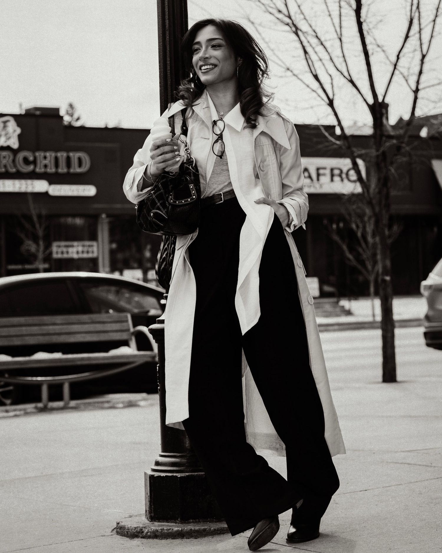Smiling woman in white trench coat and wide-leg pants poses by lamppost in black and white street photo.