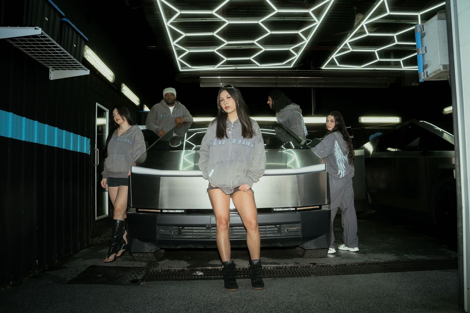 Group of four models in grey oversized hoodies posing near a car under neon lights.