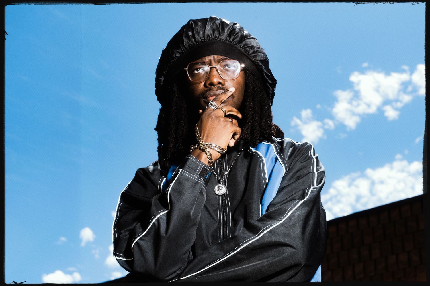 Person with dreadlocks in black tracksuit poses confidently against blue sky.