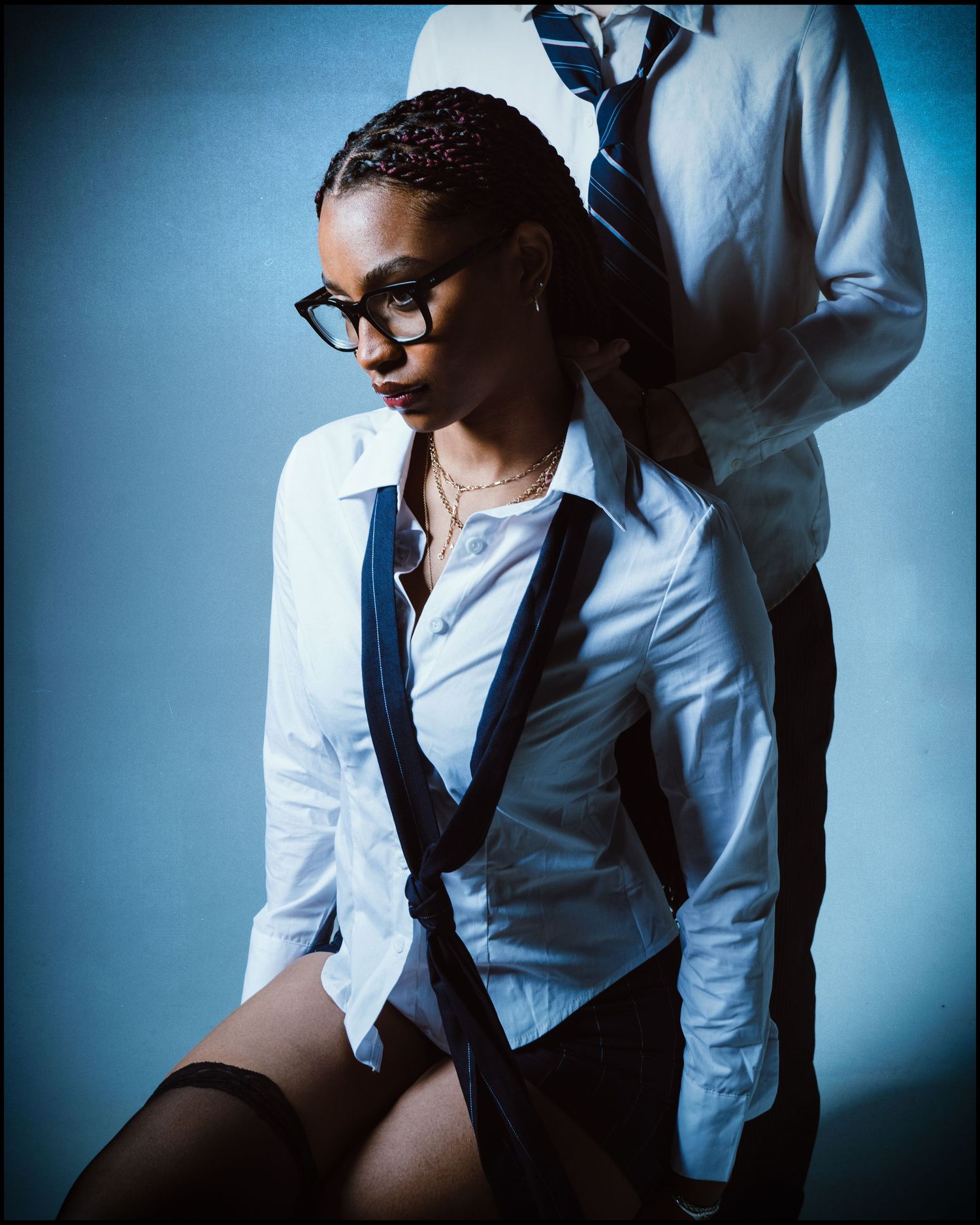 Woman in school uniform with suspenders and glasses leans against a man in a white shirt and tie.