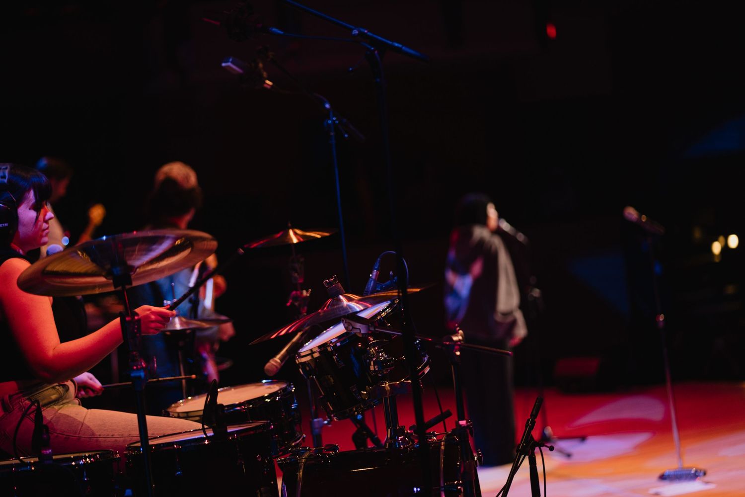 Musicians perform on a dimly lit stage with a drummer in the foreground and a singer in the background.