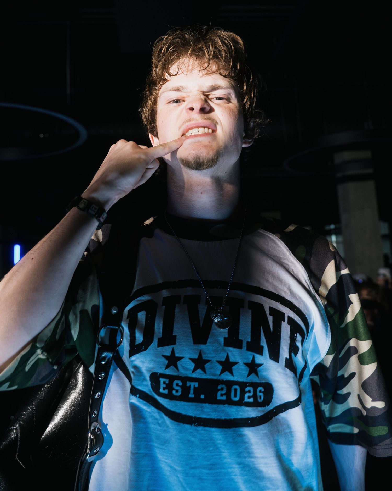 Young man in Divine EST 2026 graphic tee poses confidently at a dimly lit urban fashion event.