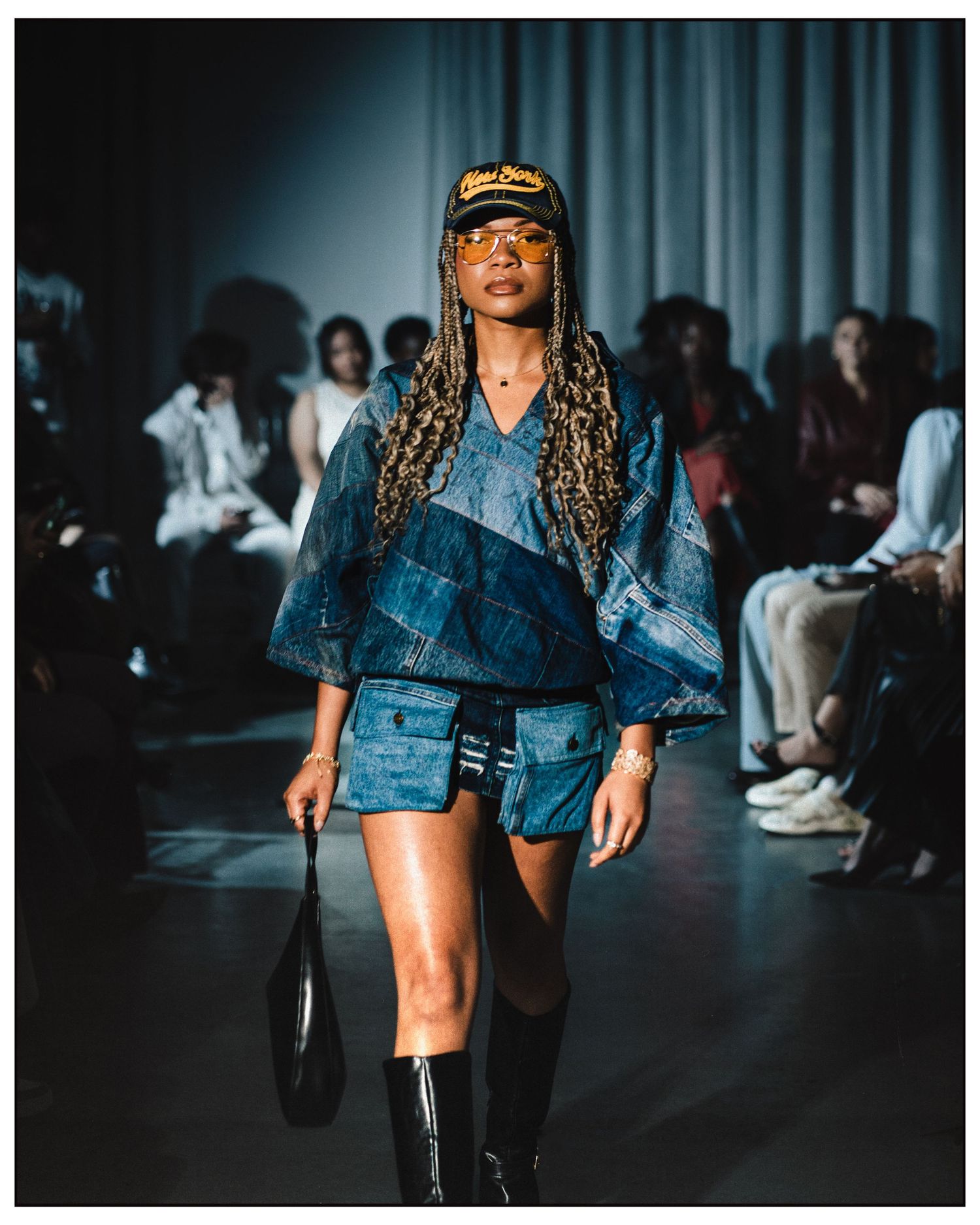 Model walks runway in denim patchwork outfit with cap, boots, and handbag at fashion show.