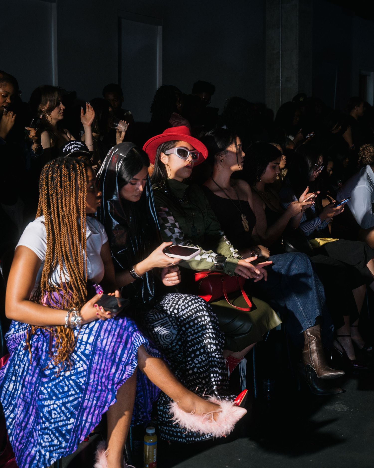 Diverse audience sits front row at indoor fashion show, with woman in red hat prominently featured.