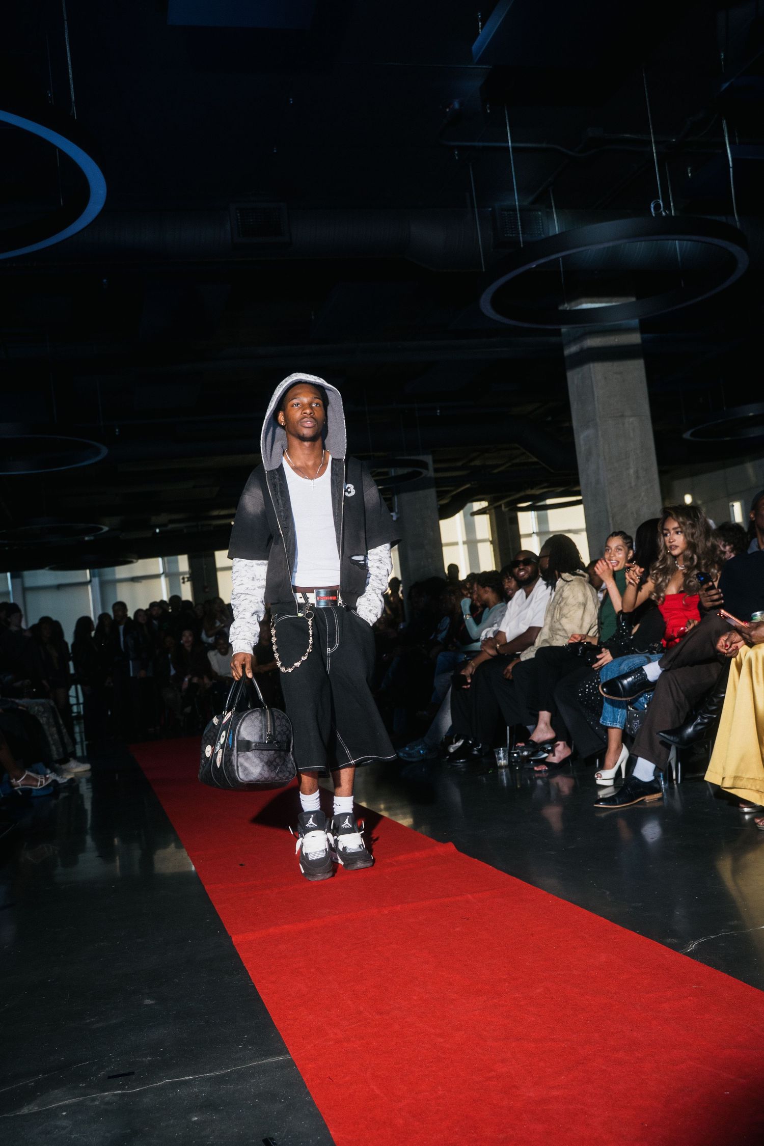 Male model in black hoodie, shorts, white socks, and Jordan sneakers carries duffle bag on runway.