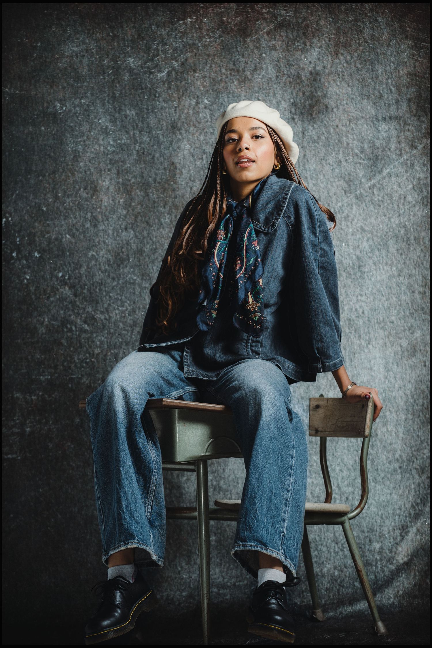 Woman in denim outfit with white beret sitting on chair against gray backdrop.