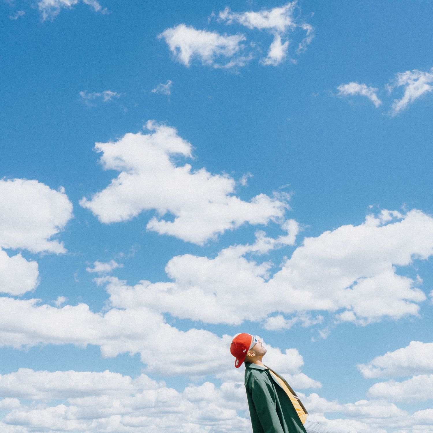 Person in red cap and green jacket looking up at a dramatic blue sky filled with white clouds.