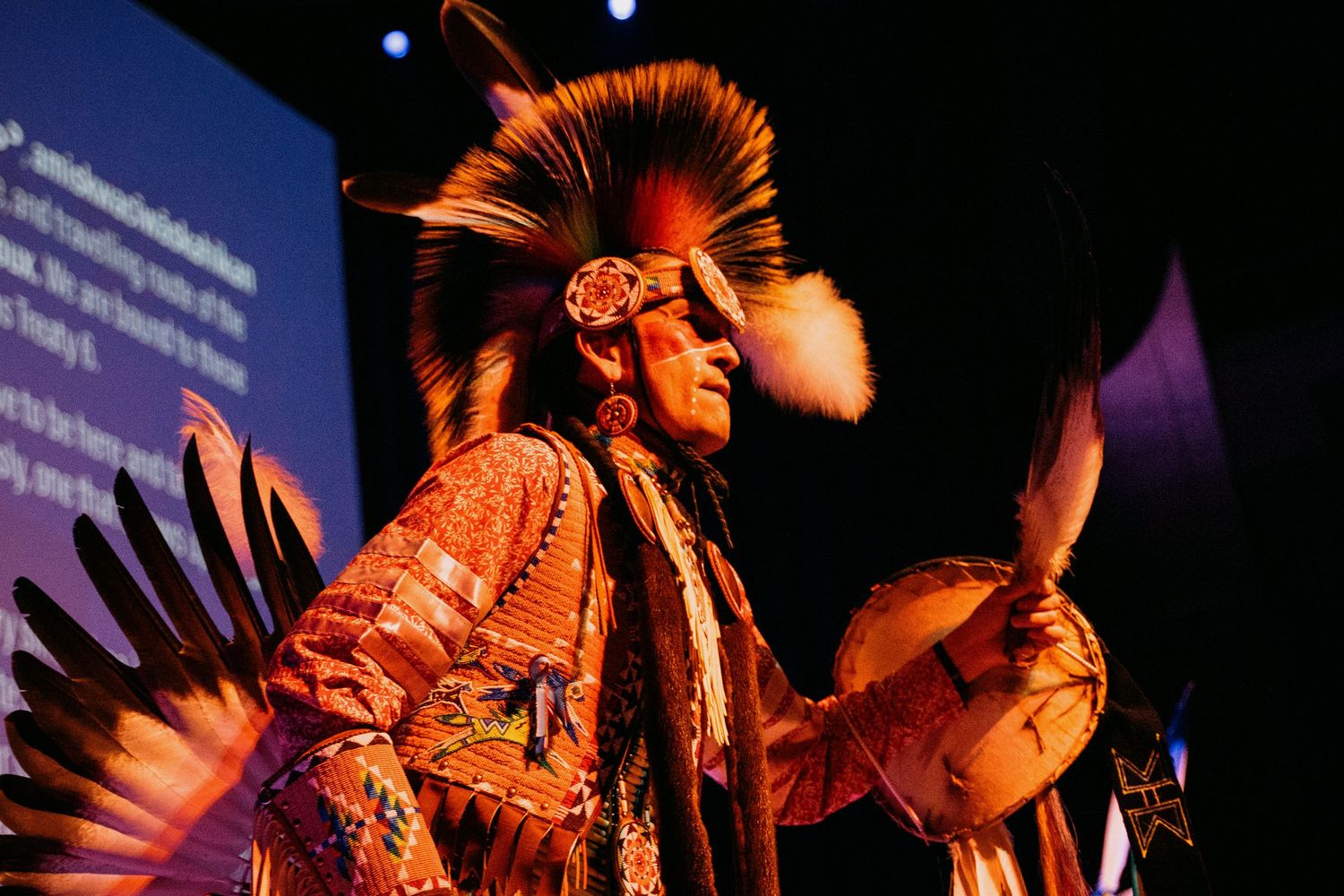 Native American dancer in traditional feathered regalia performs on a dimly lit stage at a festival.