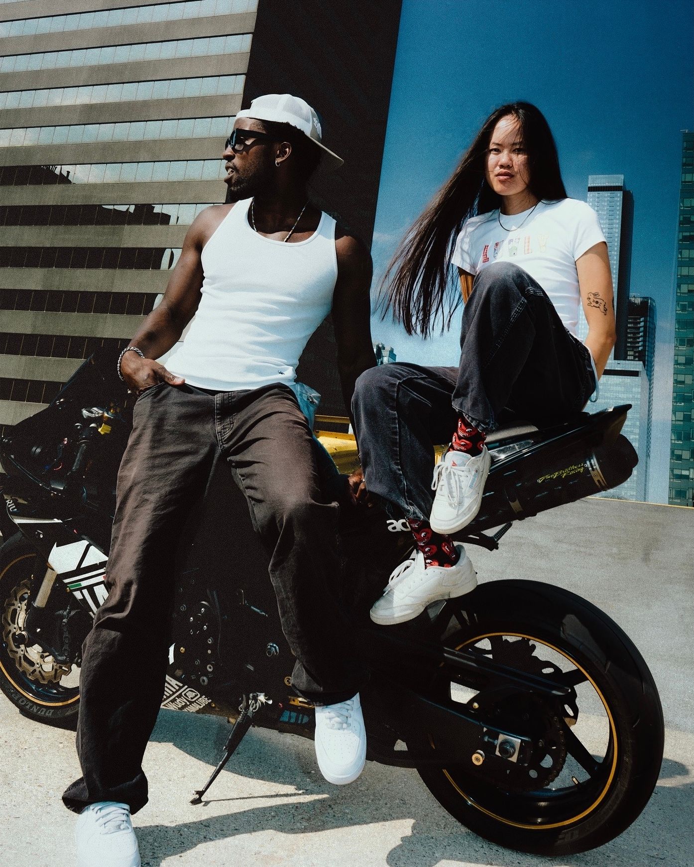 Two friends posing with a black motorcycle in front of a modern building on a sunny day.