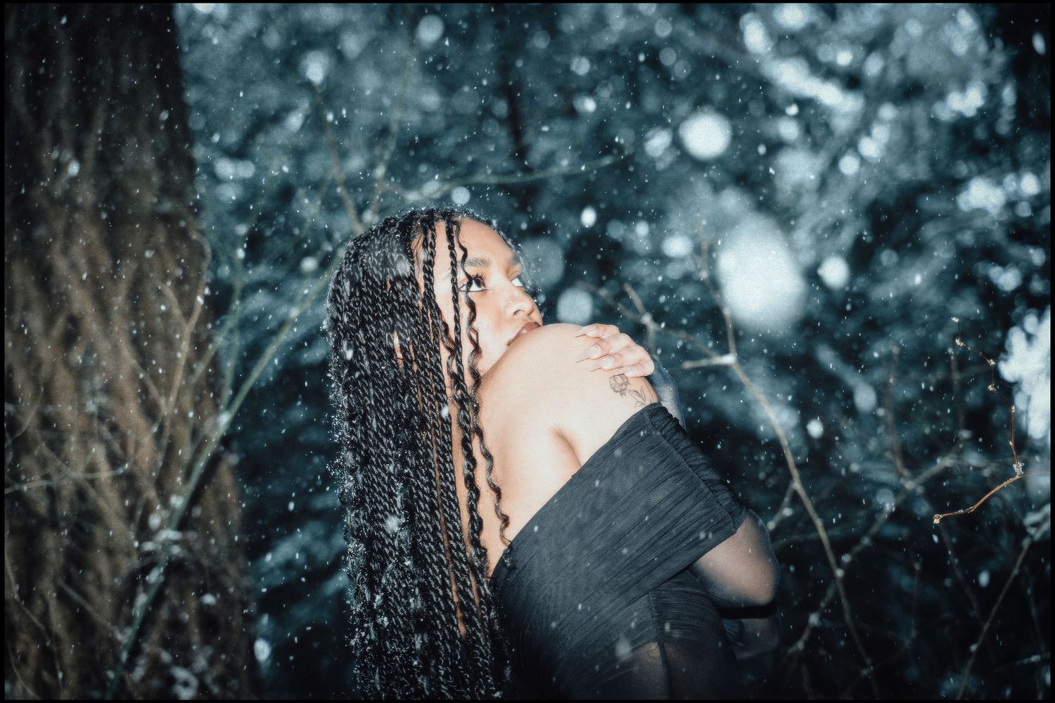 Woman with braids in black off-shoulder outfit standing in snowy forest.