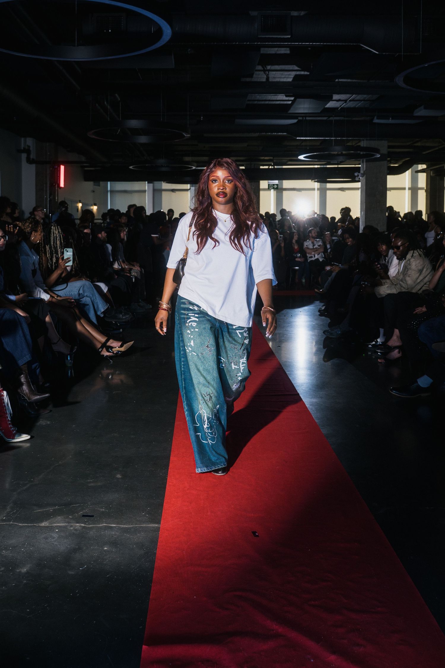 Model walks red carpet runway in white tee and wide-leg graffiti jeans at fashion show.