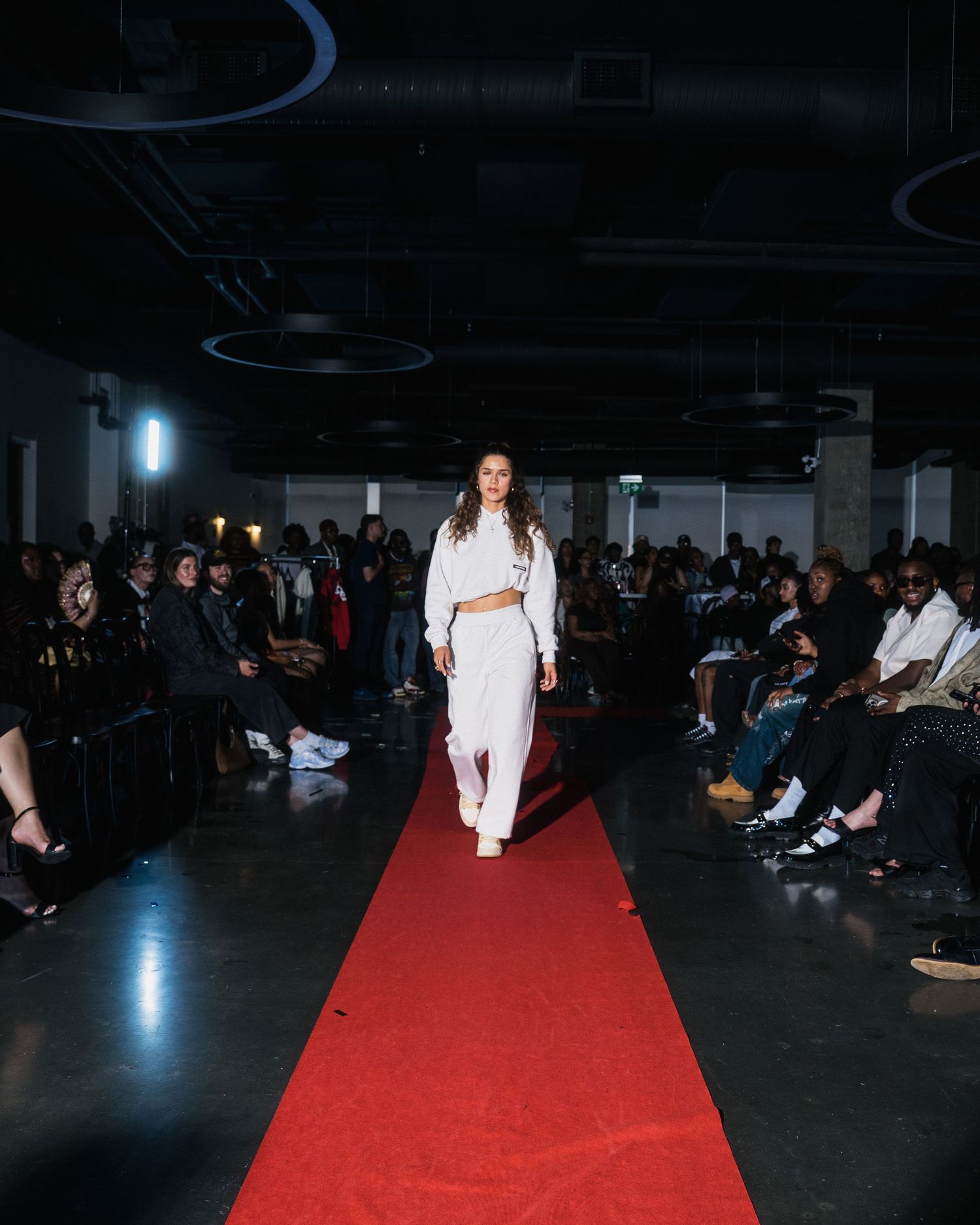 Female model in all-white cropped set walking a red carpet runway at an indoor fashion show.