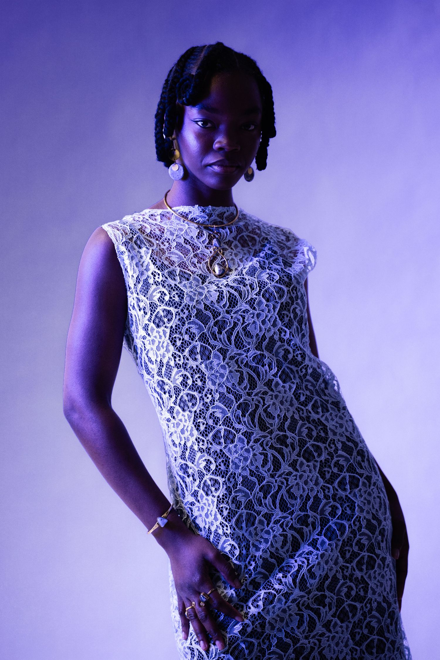 Woman in white lace dress with gold jewelry poses under purple studio lighting.
