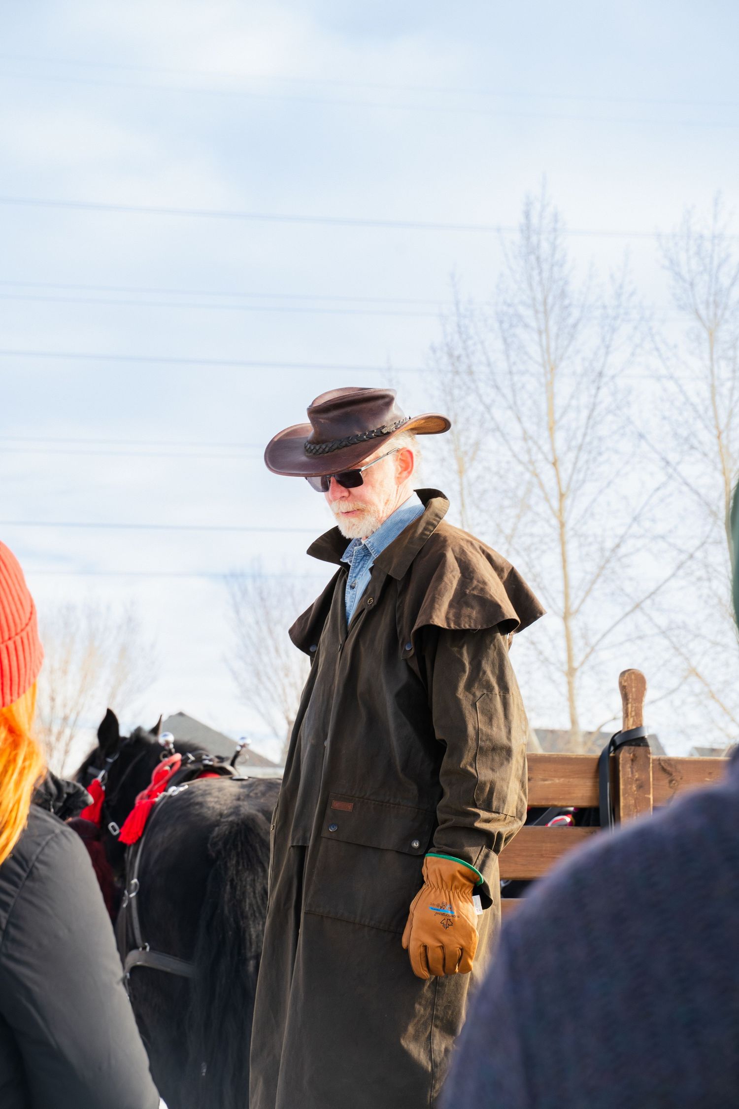 Older man in cowboy hat and long brown coat standing near horses at outdoor winter event.