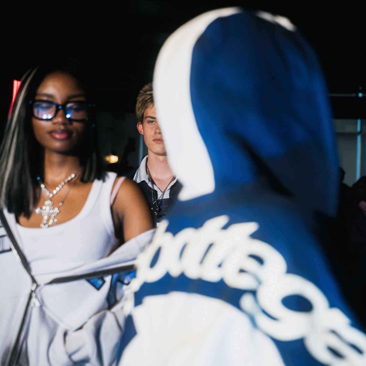 Two attendees interact with person in blue Bottega hoodie at a dimly lit fashion event.