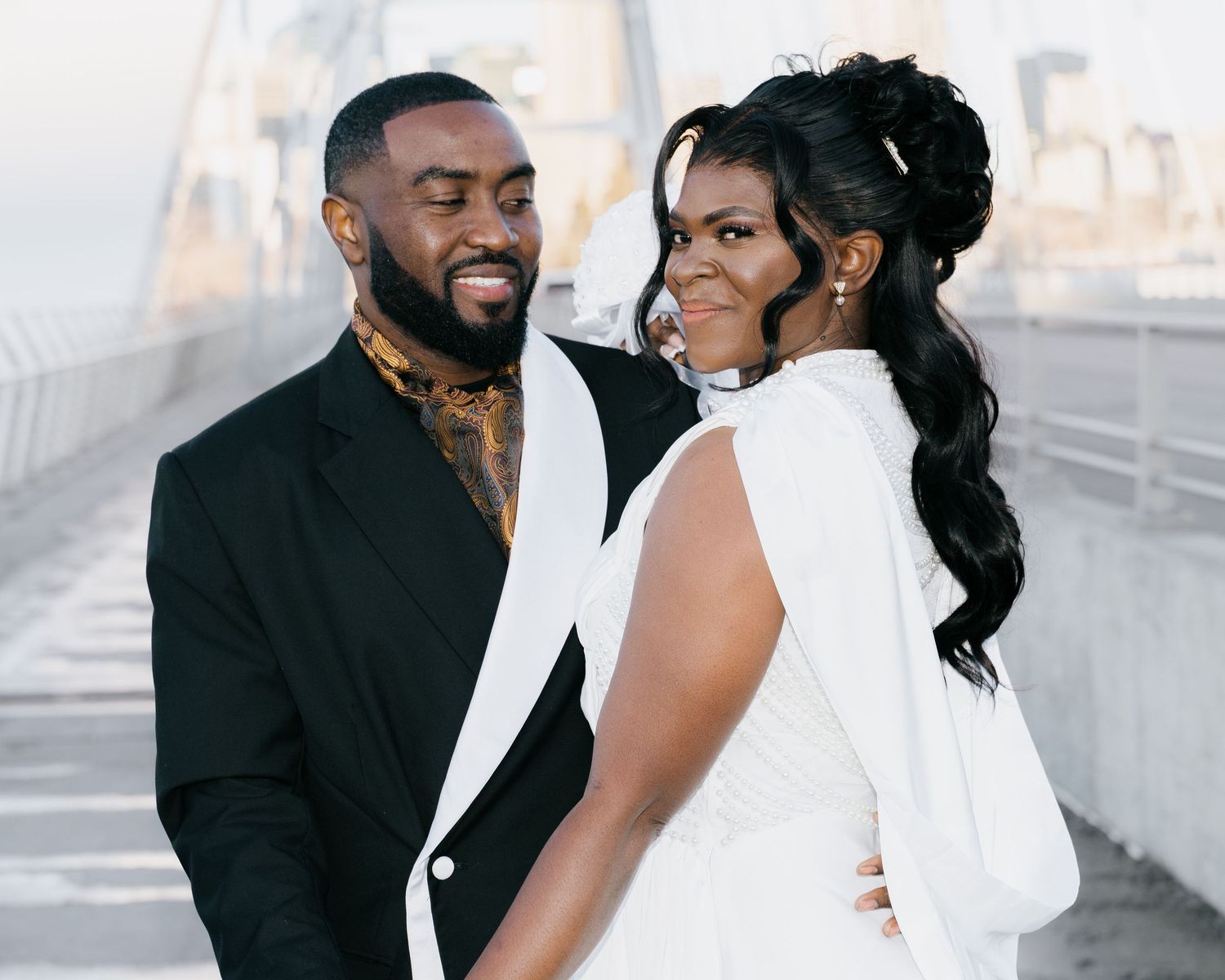 Couple in wedding attire smiling on bridge with city skyline in background.