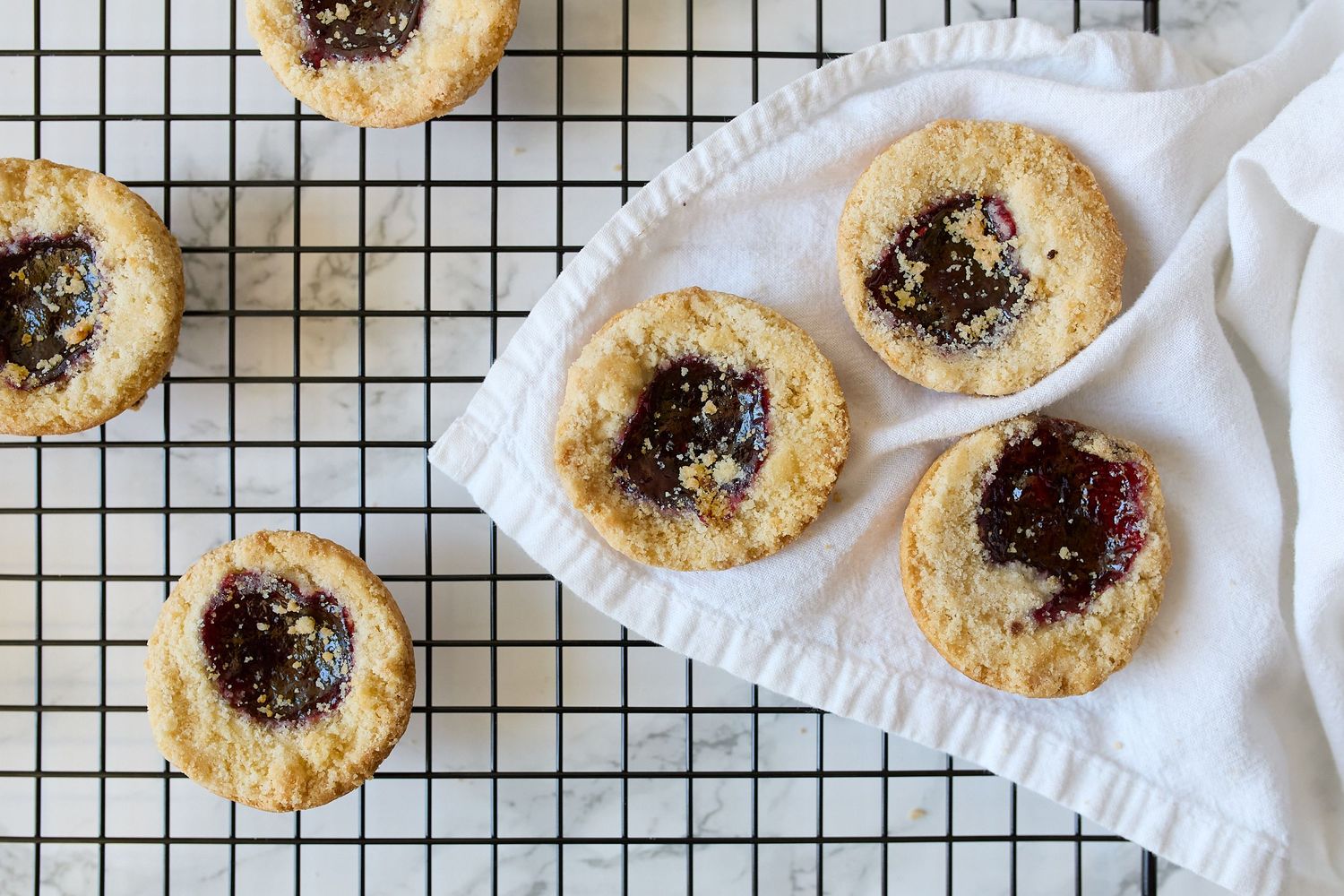 Overhead food photograph of golden jam-filled cookies cooling on a wire rack beside a white linen cloth.