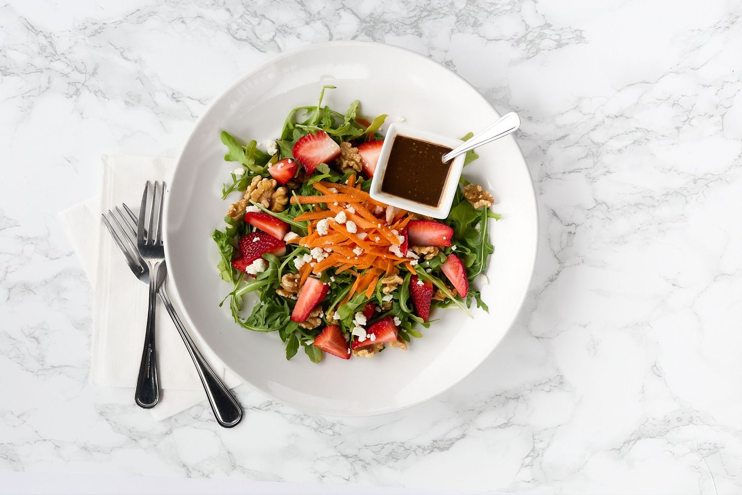 Overhead food photograph of fresh arugula salad with strawberries, shredded carrots, and dressing on a white plate against a marble surface.