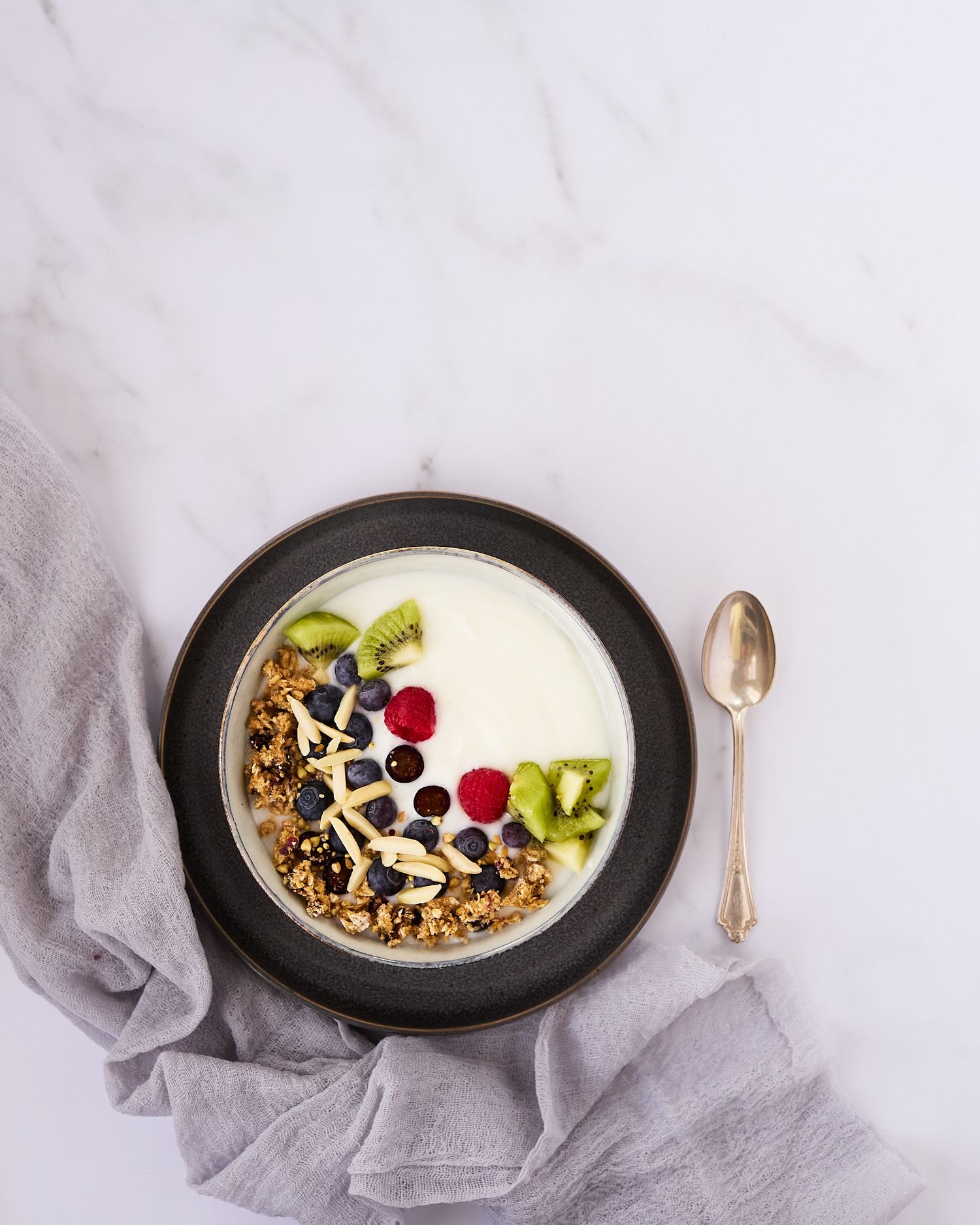 Overhead food photograph of a yogurt bowl topped with granola, fresh berries, kiwi, and nuts on a pale marble surface.