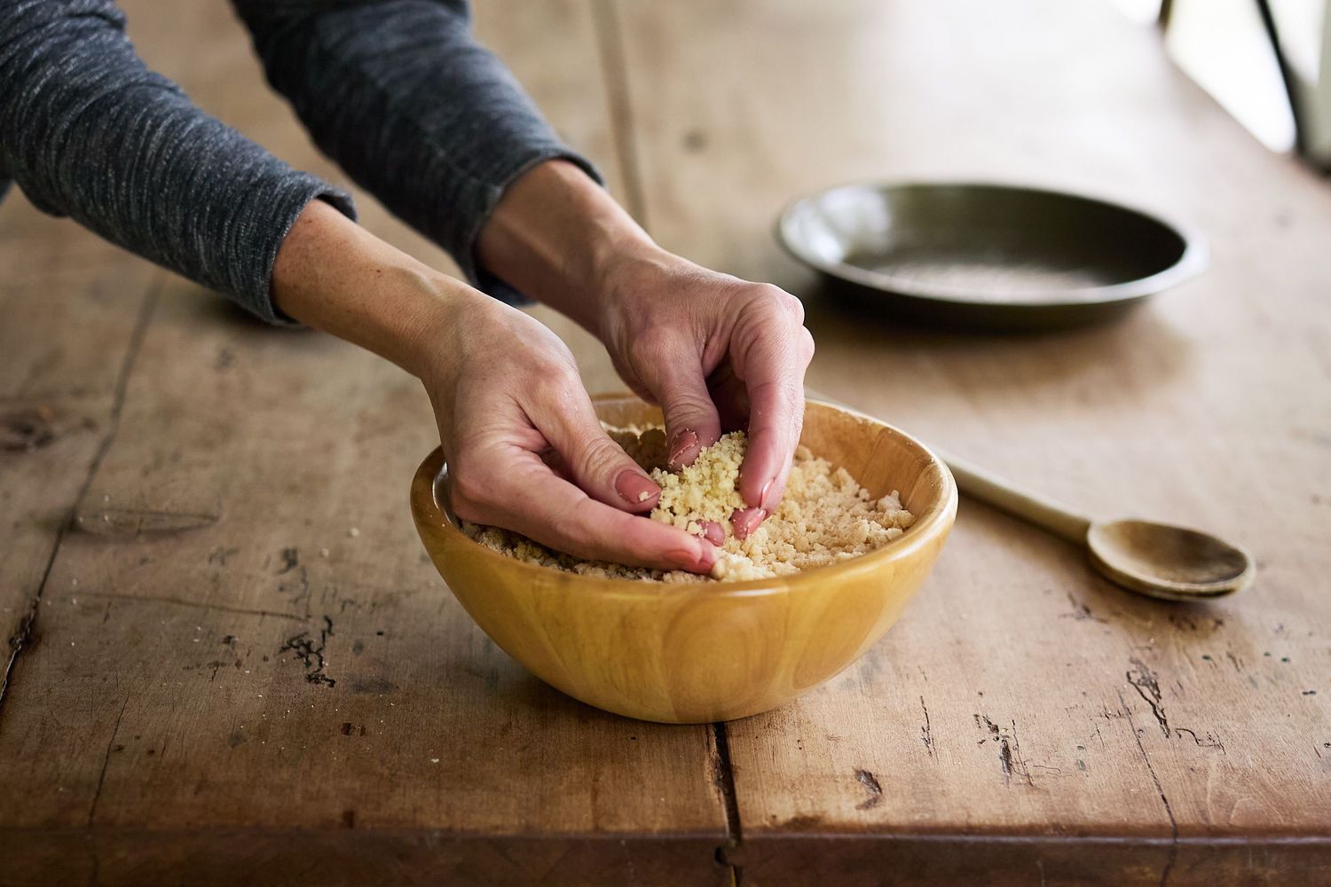 Hands preparing a homemade Pie Provisions pie crust in a natural wood bowl on a rustic wooden work surface.