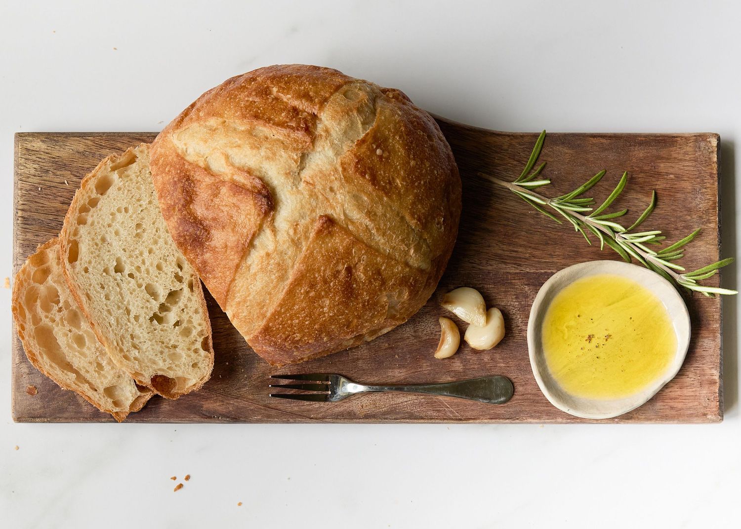 Rustic still life of a crusty sourdough loaf with a slice of bread, small bowl of TruTruffle oil , garlic, and rosemary on a wooden board.