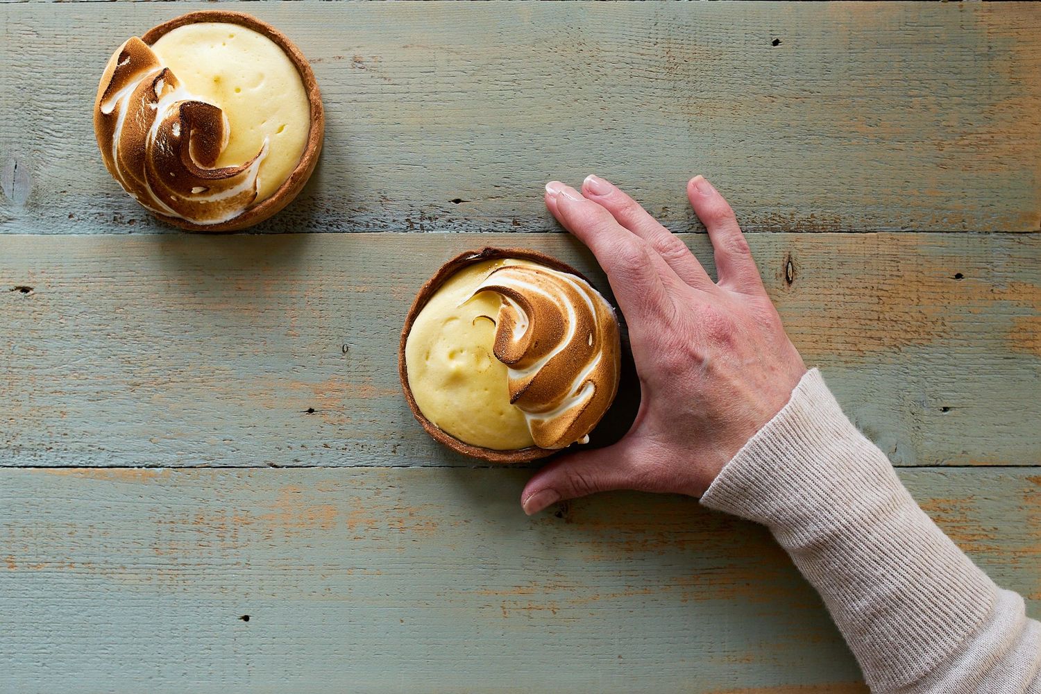 Hands arranging a pastry topped with cream on a weathered blue tabletop, photographed from above.