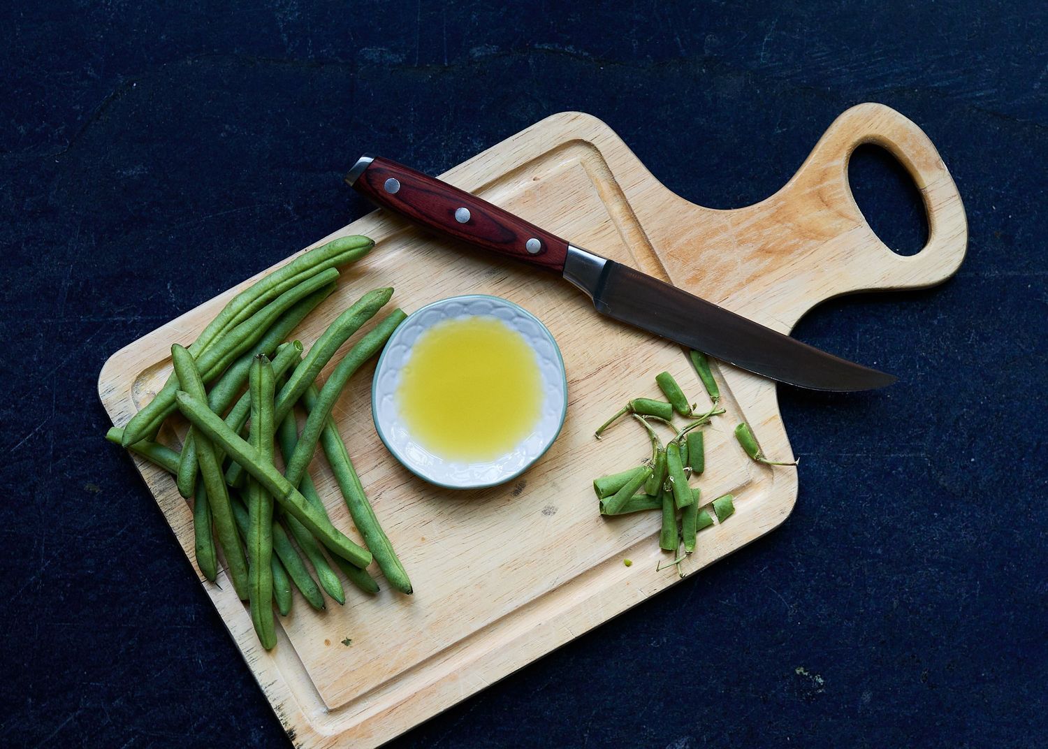 Rustic food still life of fresh green beans, TruTruffle oil, and a chef’s knife on a wooden cutting board against a dark background.