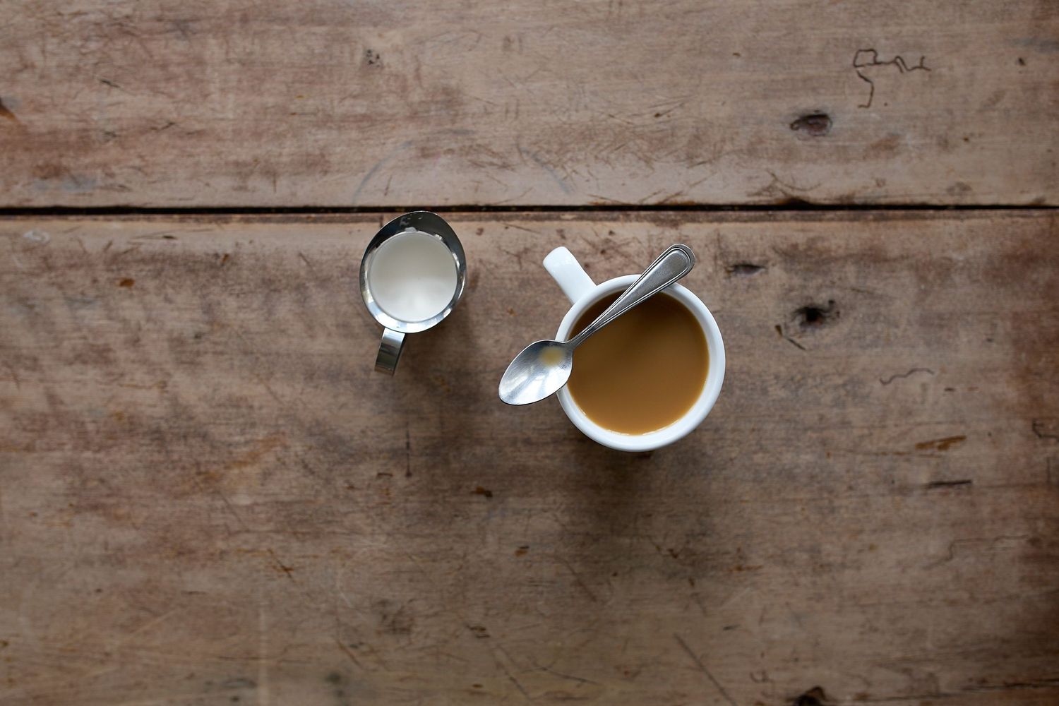Minimalist overhead still life of a cup of coffee, a small glass of cream, and a spoon on a rustic wooden tabletop.