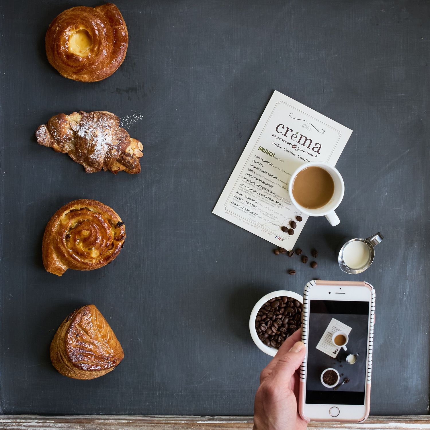 Overhead editorial food photograph of golden pastries, coffee, and a smartphone on a dark blue surface, with a hand capturing the scene.