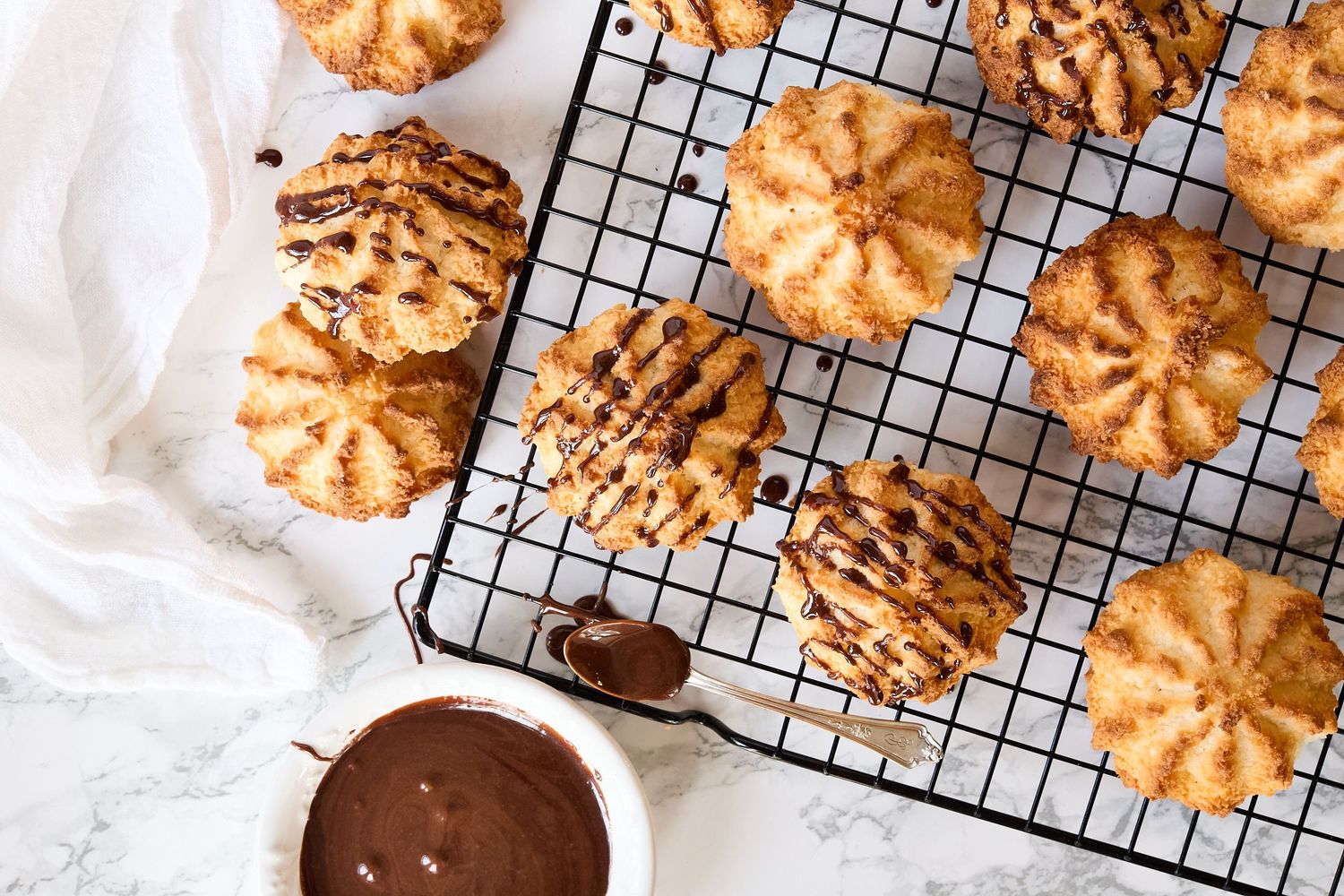 Assortment of freshly baked coconut cookies drizzled with melted chocoloate, cooling on a wire rack, photographed from above.