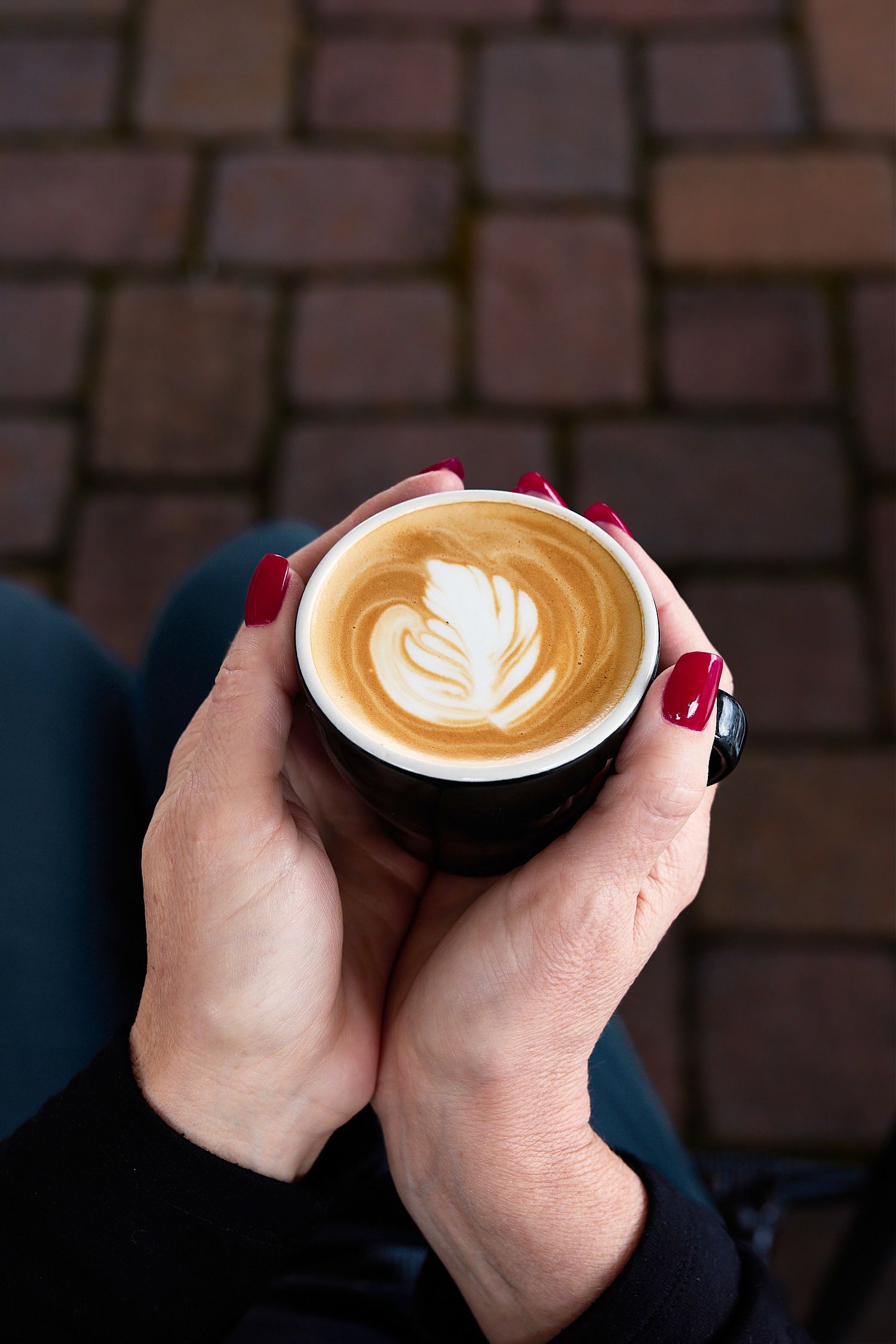 Close-up of hands holding a cappuccino with heart-shaped latte art against a brick-paved background.