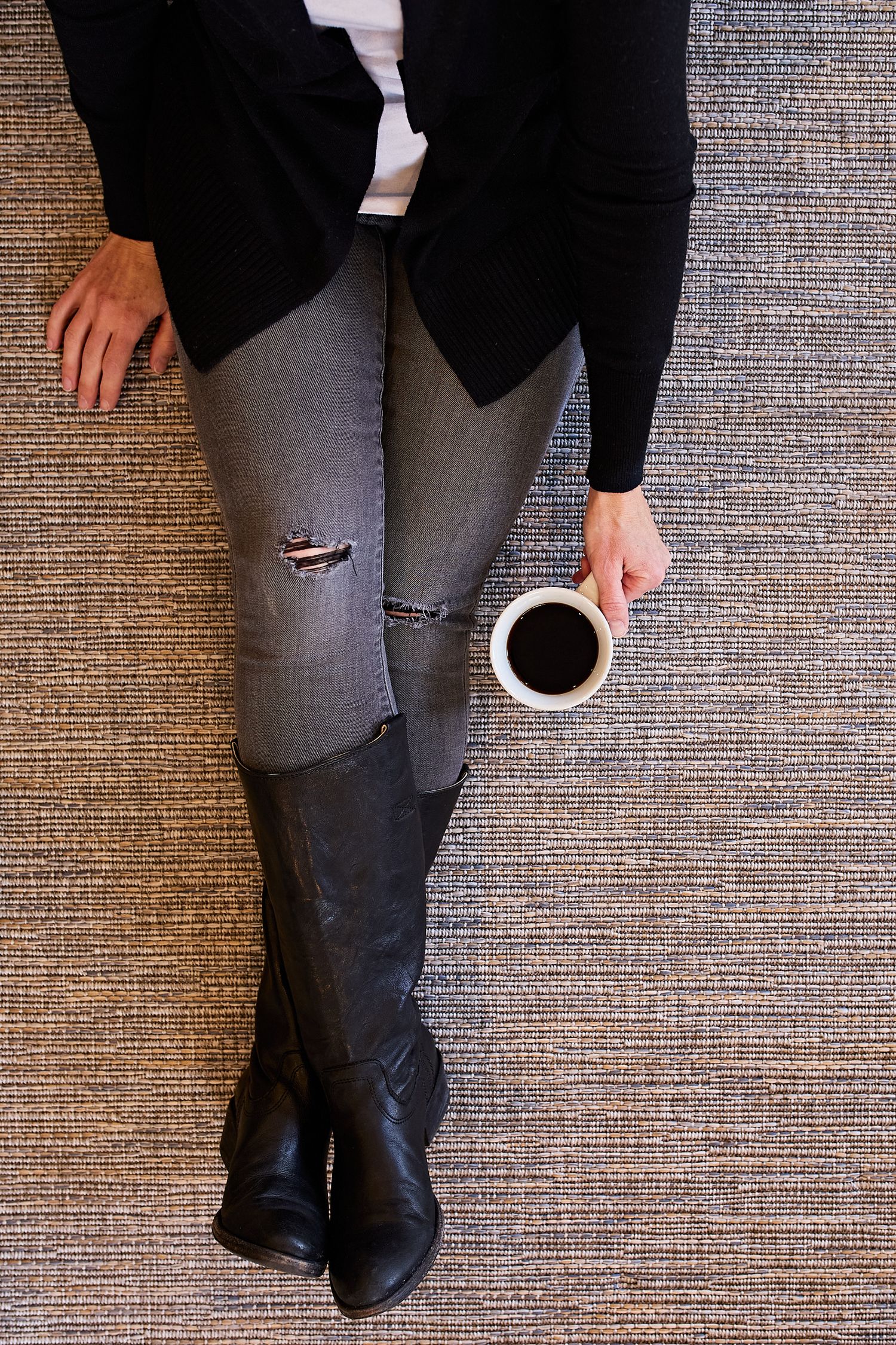 Lifestyle photograph of a woman in ripped gray jeans and black boots holding a cup of coffee while seated on a textured rug.