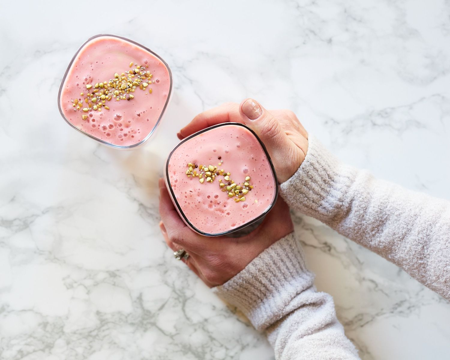 Overhead photograph of two pink smoothies topped with chopped pistachios, one held in hands above a white marble surface.