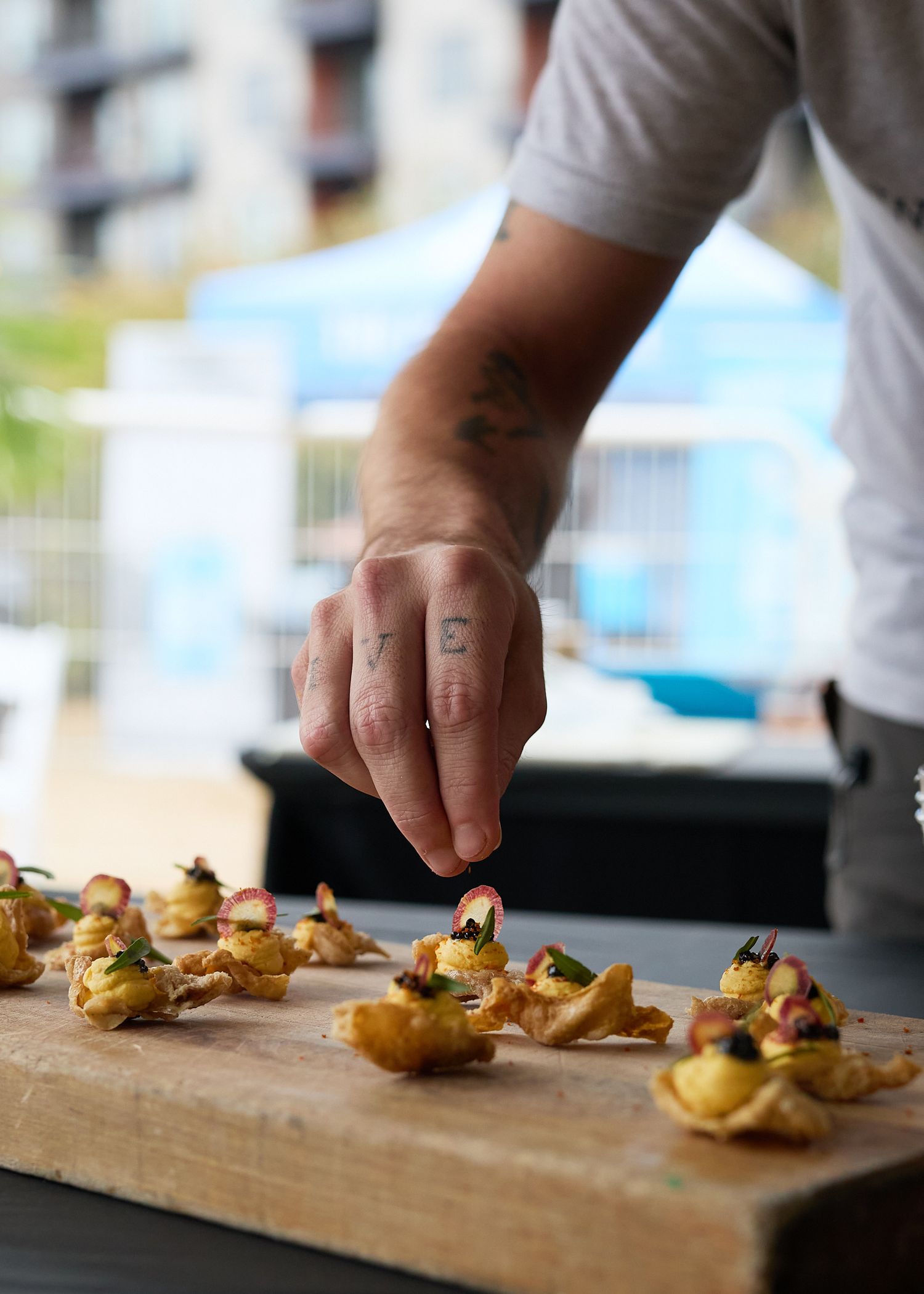 Candid culinary event photograph of a chef garnishing bite-sized appetizers arranged on a wooden serving board.