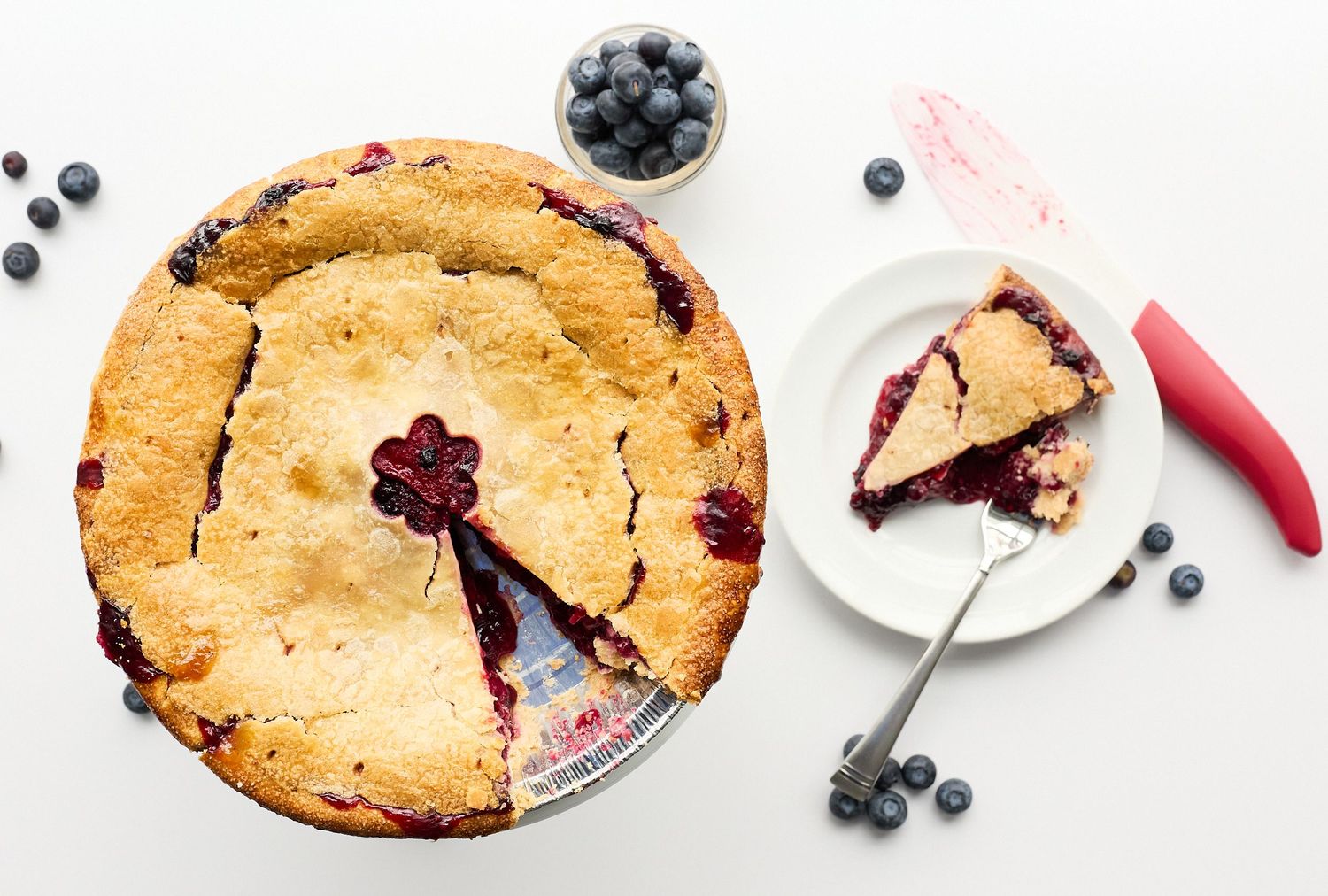 Overhead photograph of a golden blueberry pie with a slice served on a plate, fresh blueberries, and a red pie server.