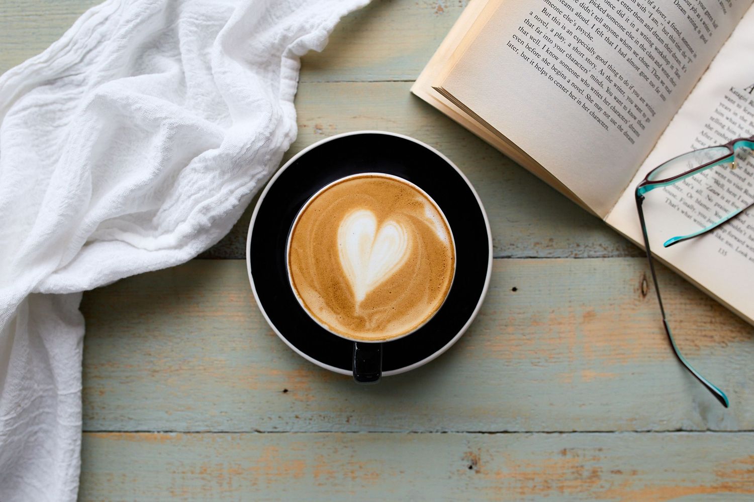 Cup of coffee with heart-shaped latte art arranged beside an open book and eyeglasses on a rustic tabletop.