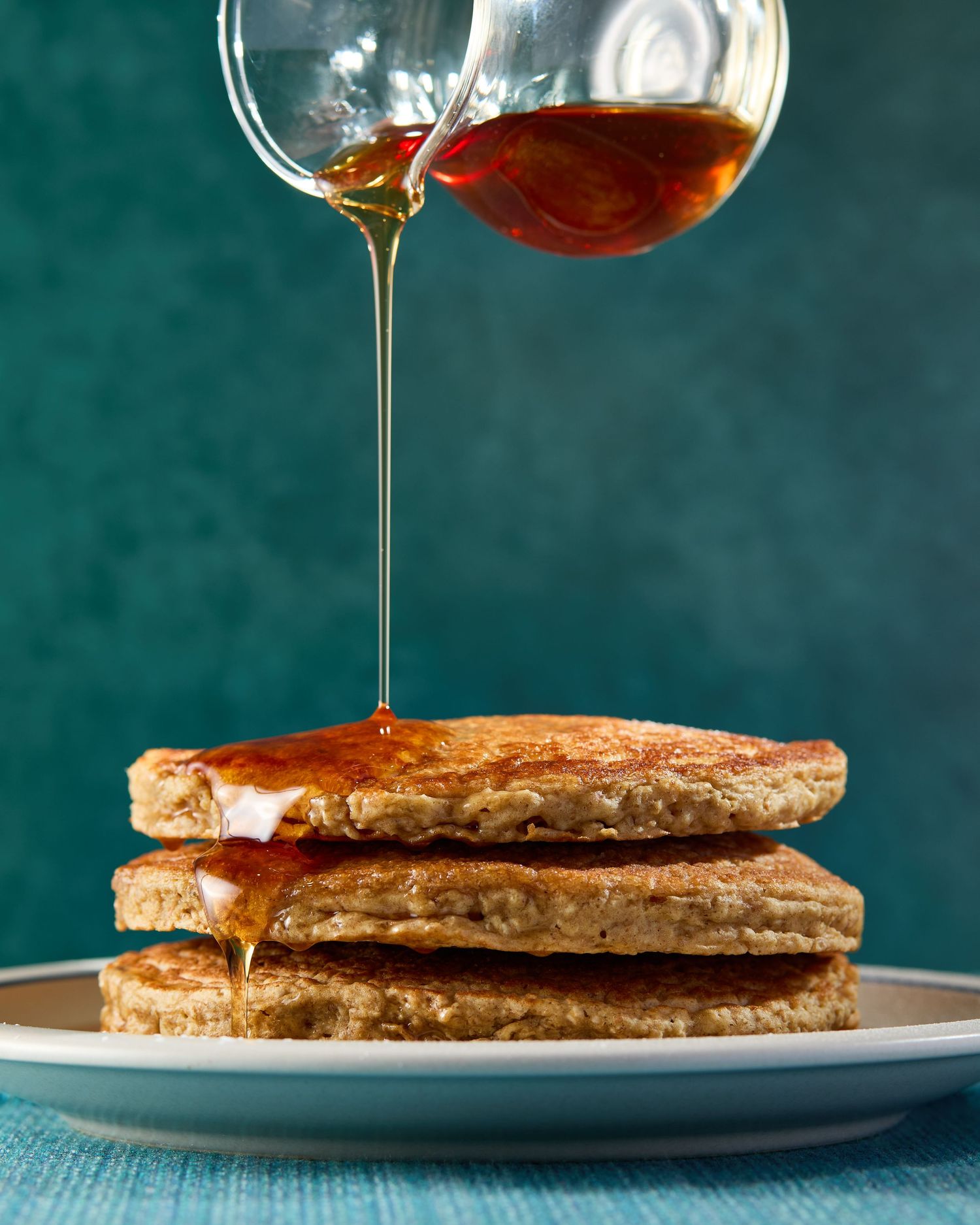 Close-up food photograph of maple syrup pouring over a stack of golden GoodGate Foods Harvest pancakes against a deep teal background.