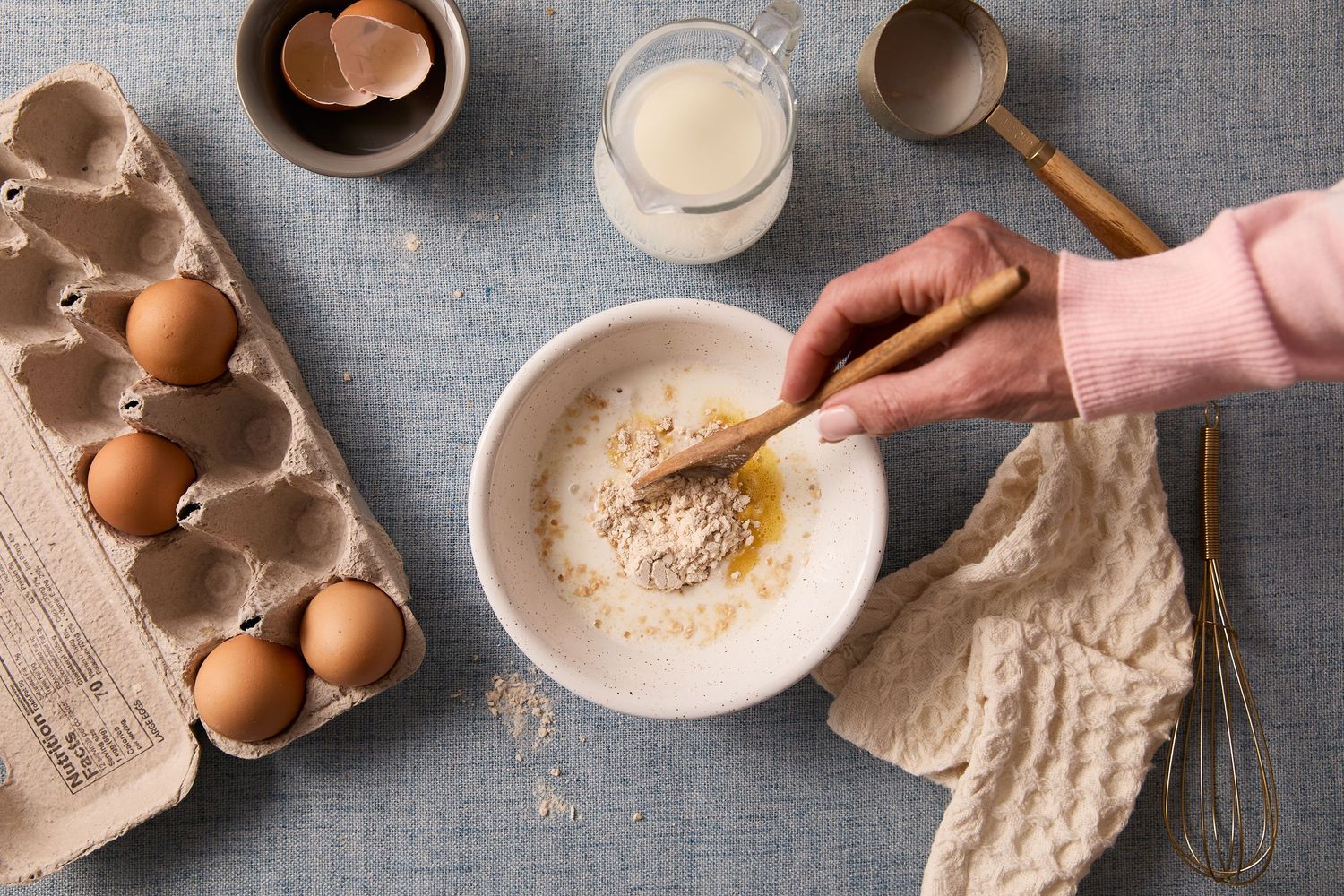 Overhead baking photograph of a hand whisking pancake batter in a white bowl beside eggs, milk, and baking utensils.