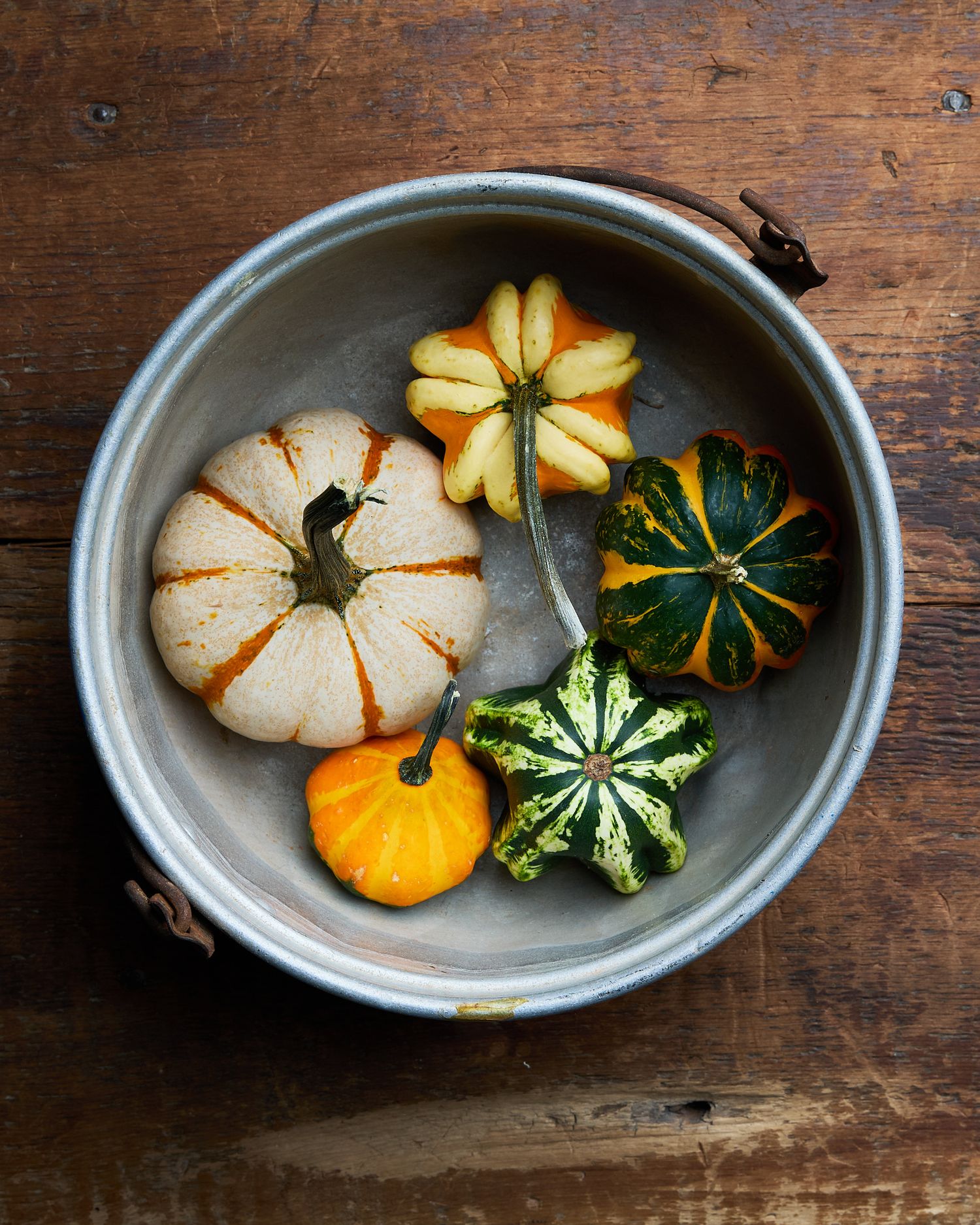 Overhead rustic still life of miniature white, yellow, and green striped squash arranged in a metal bowl on a weathered wooden table.