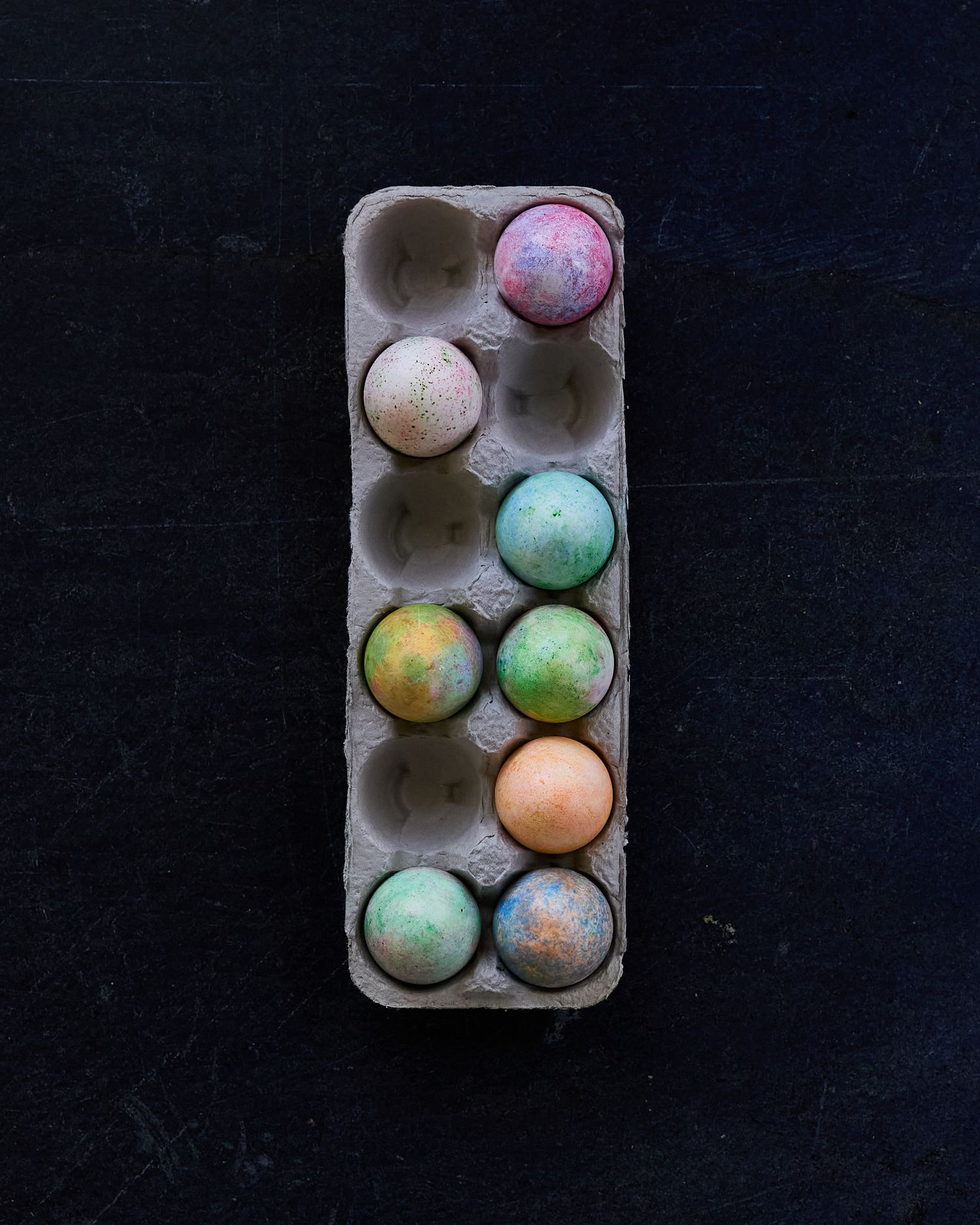 Overhead still life of pastel-colored Easter eggs in a recycled paper carton against a dark background.