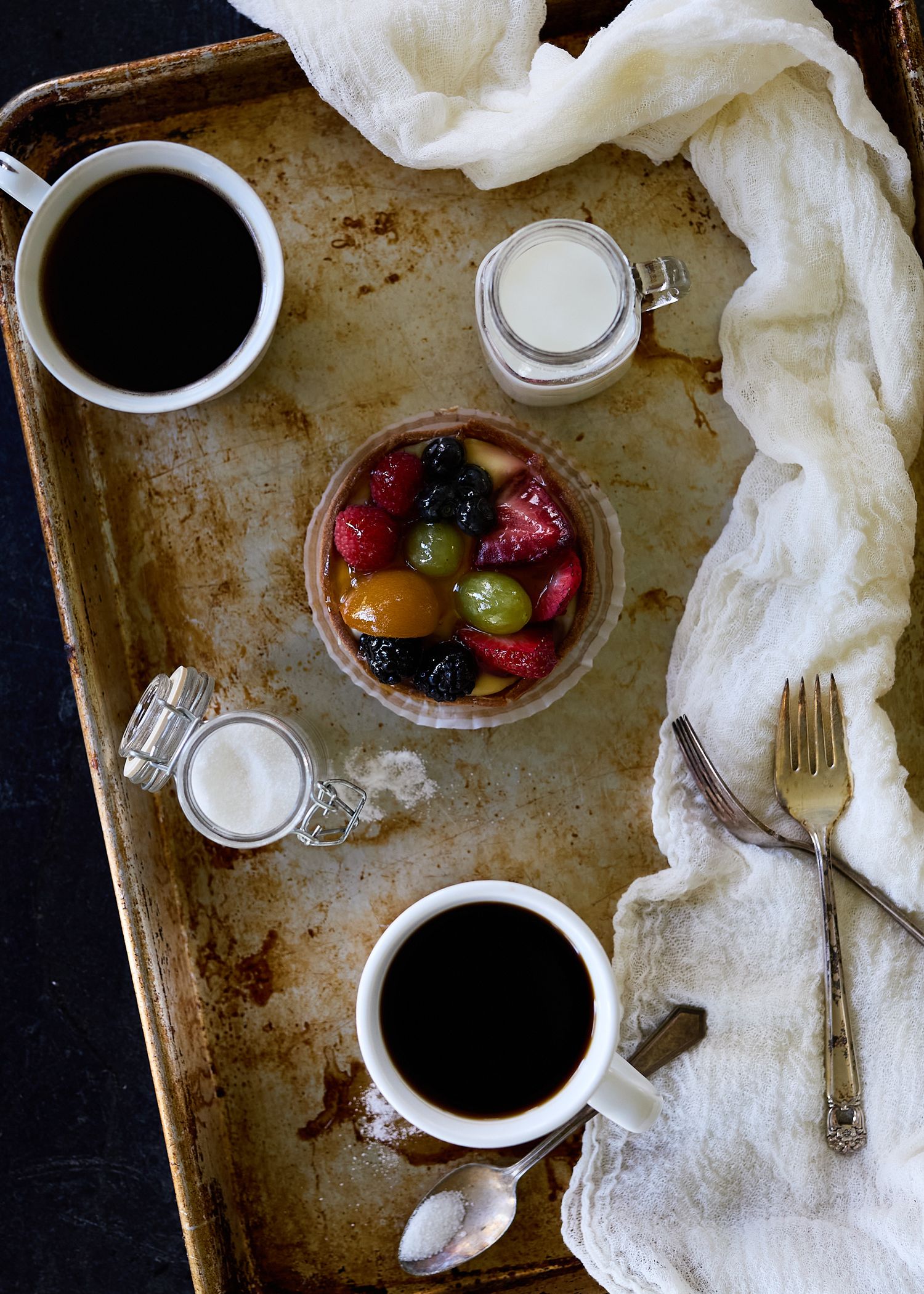 Rustic overhead still life of colorful fruit in a bowl, two cups of black coffee, glass milk jars, and linen on a weathered tray.