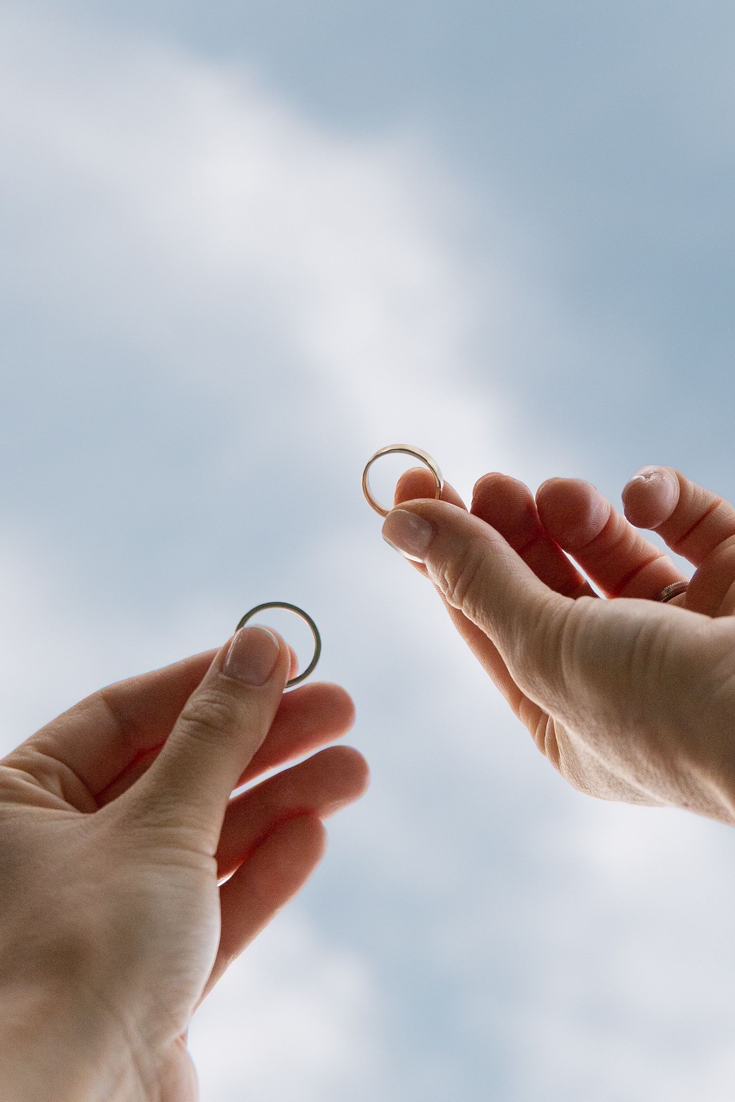 Two hands holding gold wedding rings up against a soft blue cloudy sky.