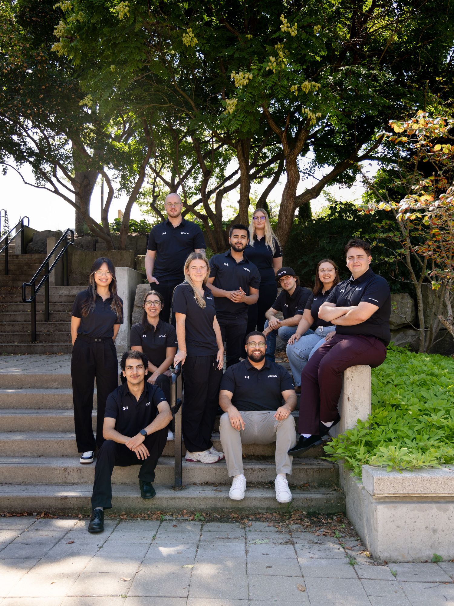 Group of ten professionals in black uniforms pose together on outdoor steps surrounded by trees.