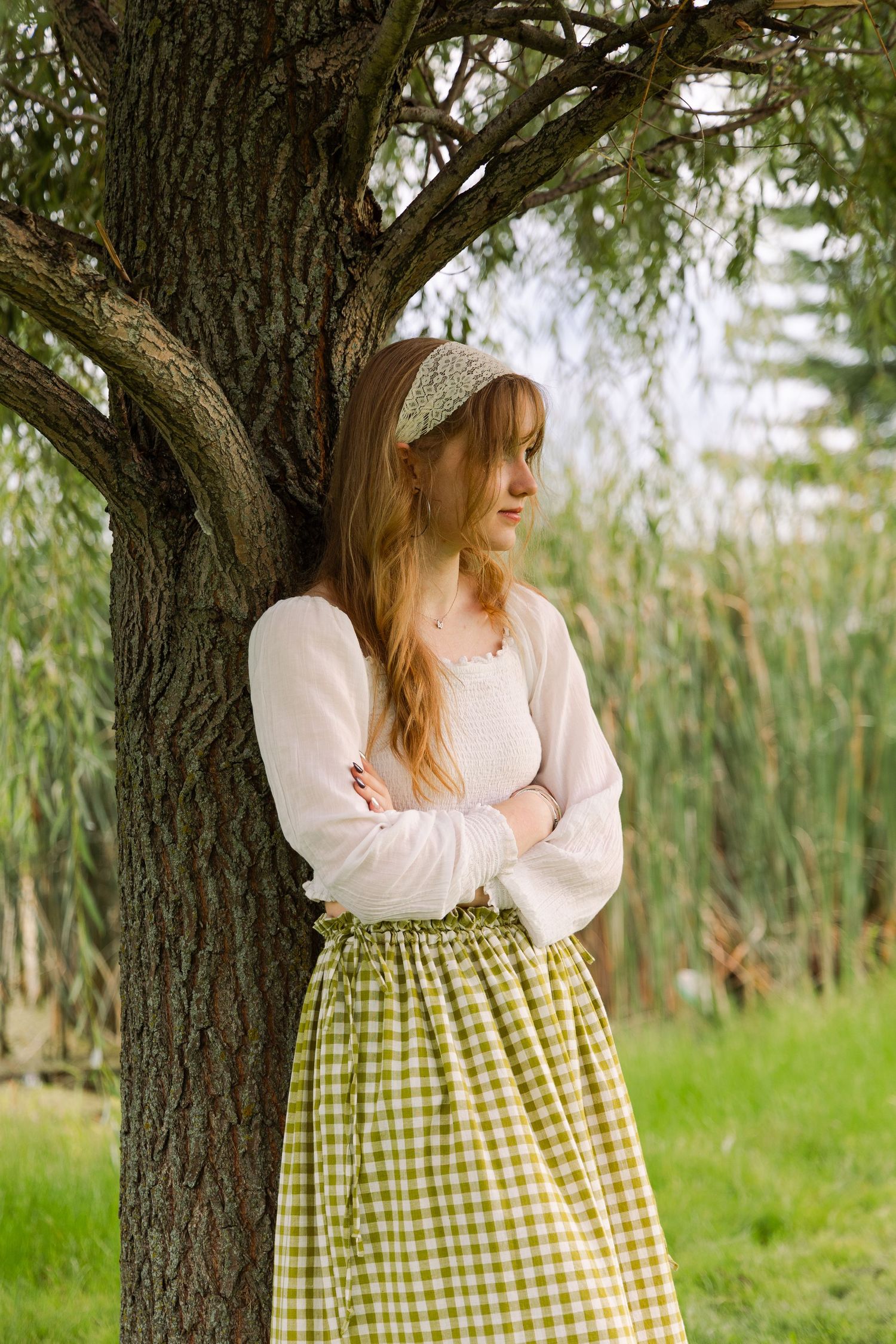 A young woman in a green gingham midi skirt and white top leans against a tree with arms crossed outdoors.
