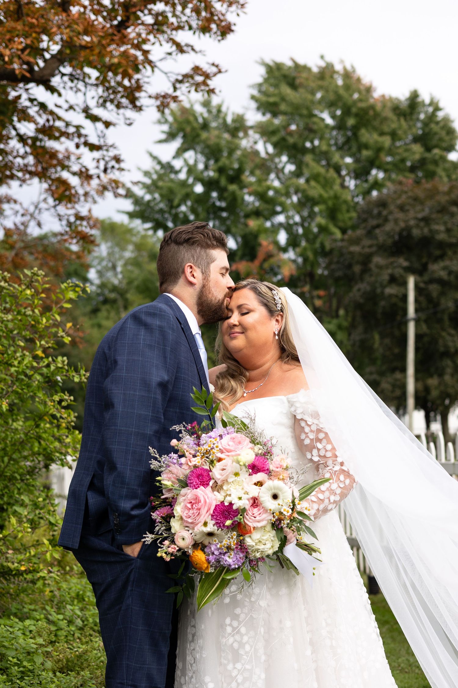 Groom in navy suit kisses bride in lace gown holding vibrant wildflower bouquet outdoors.