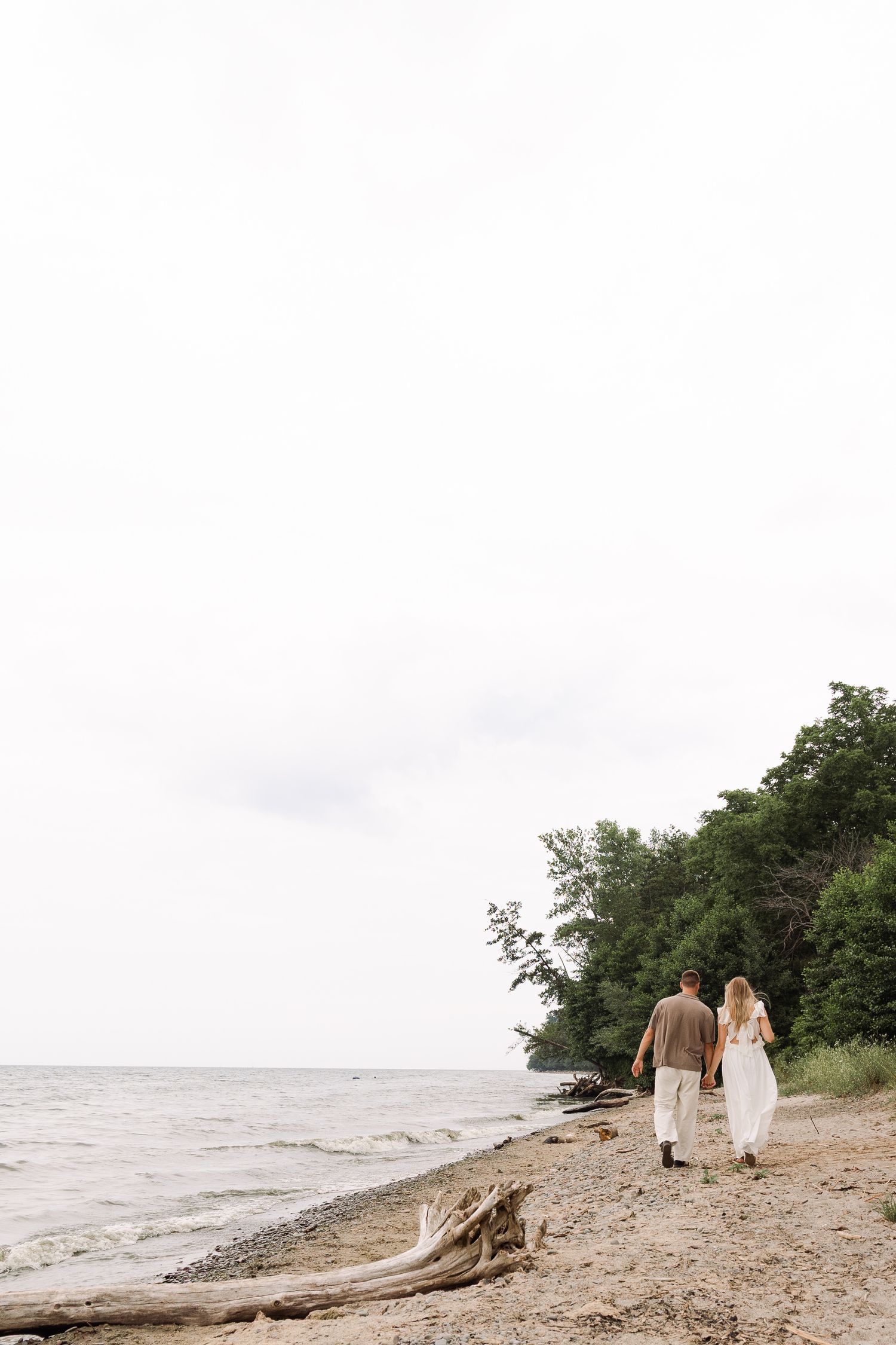Couple walking hand in hand along a rocky shoreline with green trees and overcast sky.