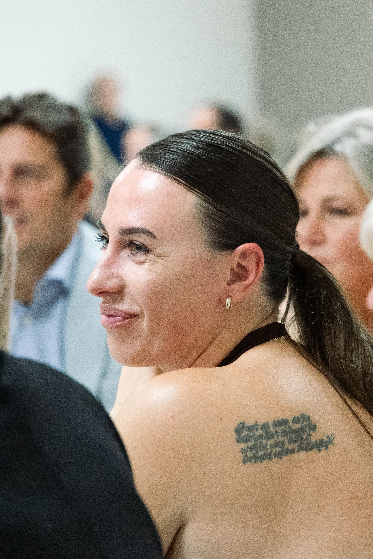 Woman with dark hair in ponytail and script tattoo on shoulder smiling at an indoor wedding reception.