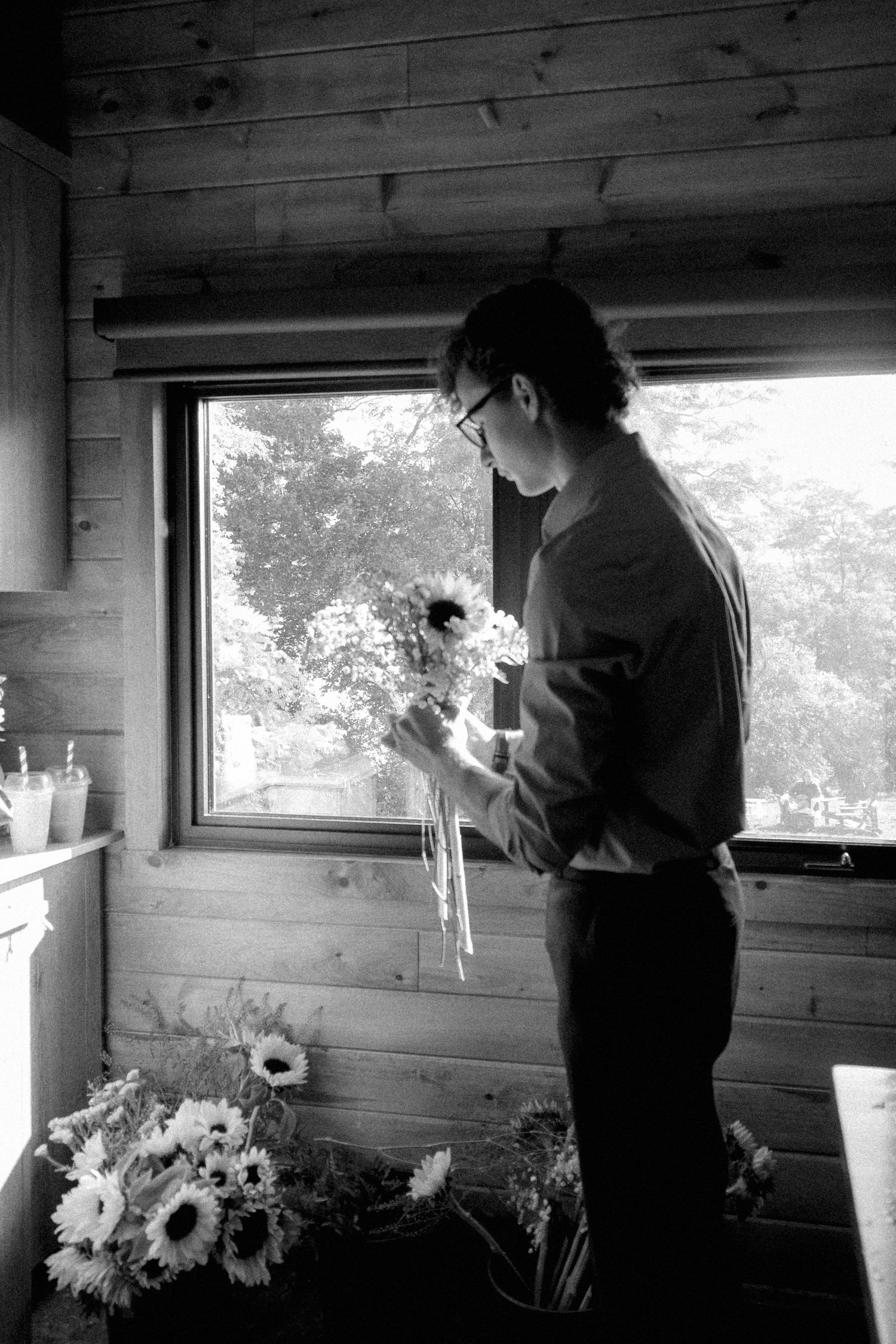 Black and white photo of man arranging sunflowers by cabin window with natural light streaming in.