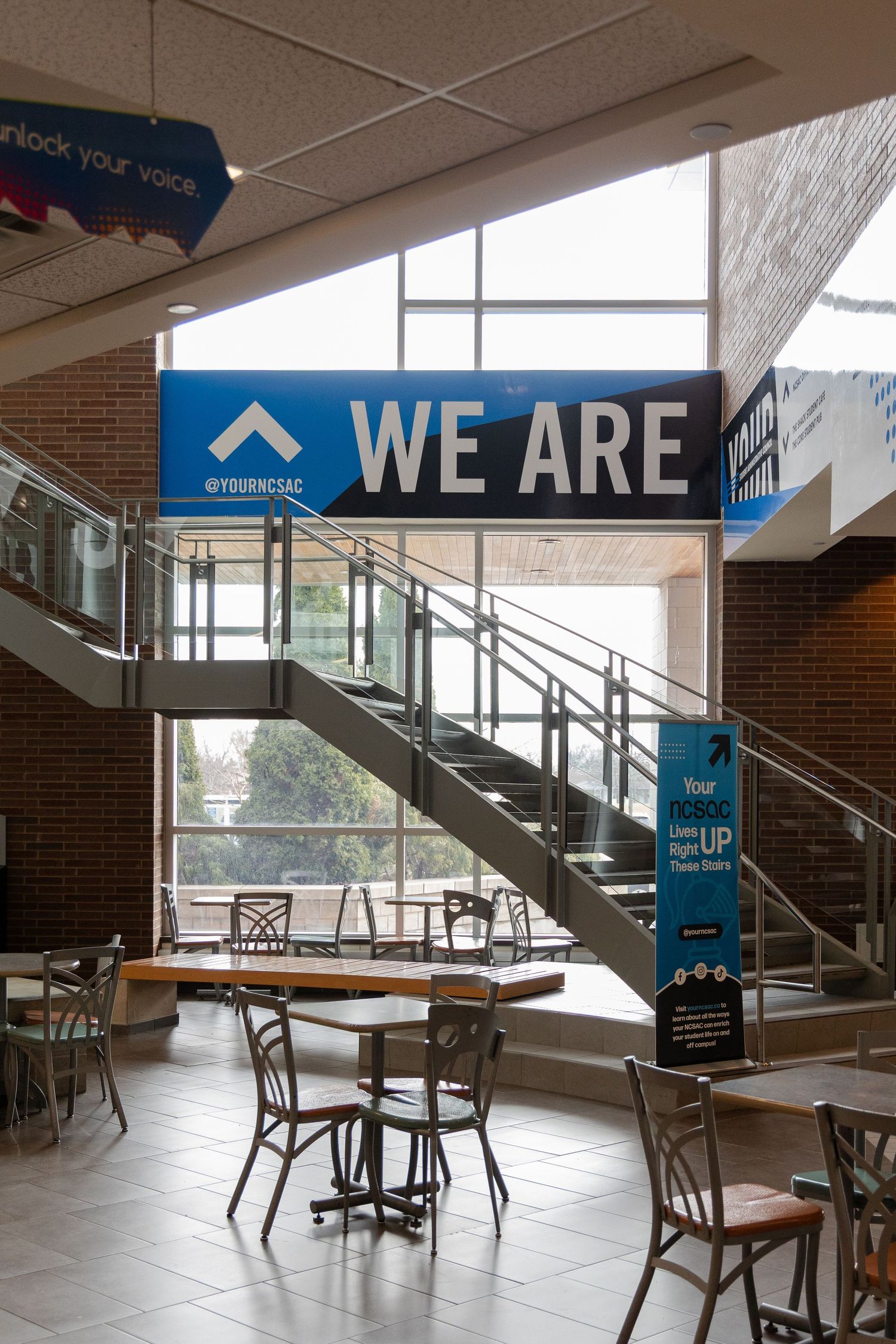 Modern university student center interior with blue WE ARE sign above staircase and seating area.