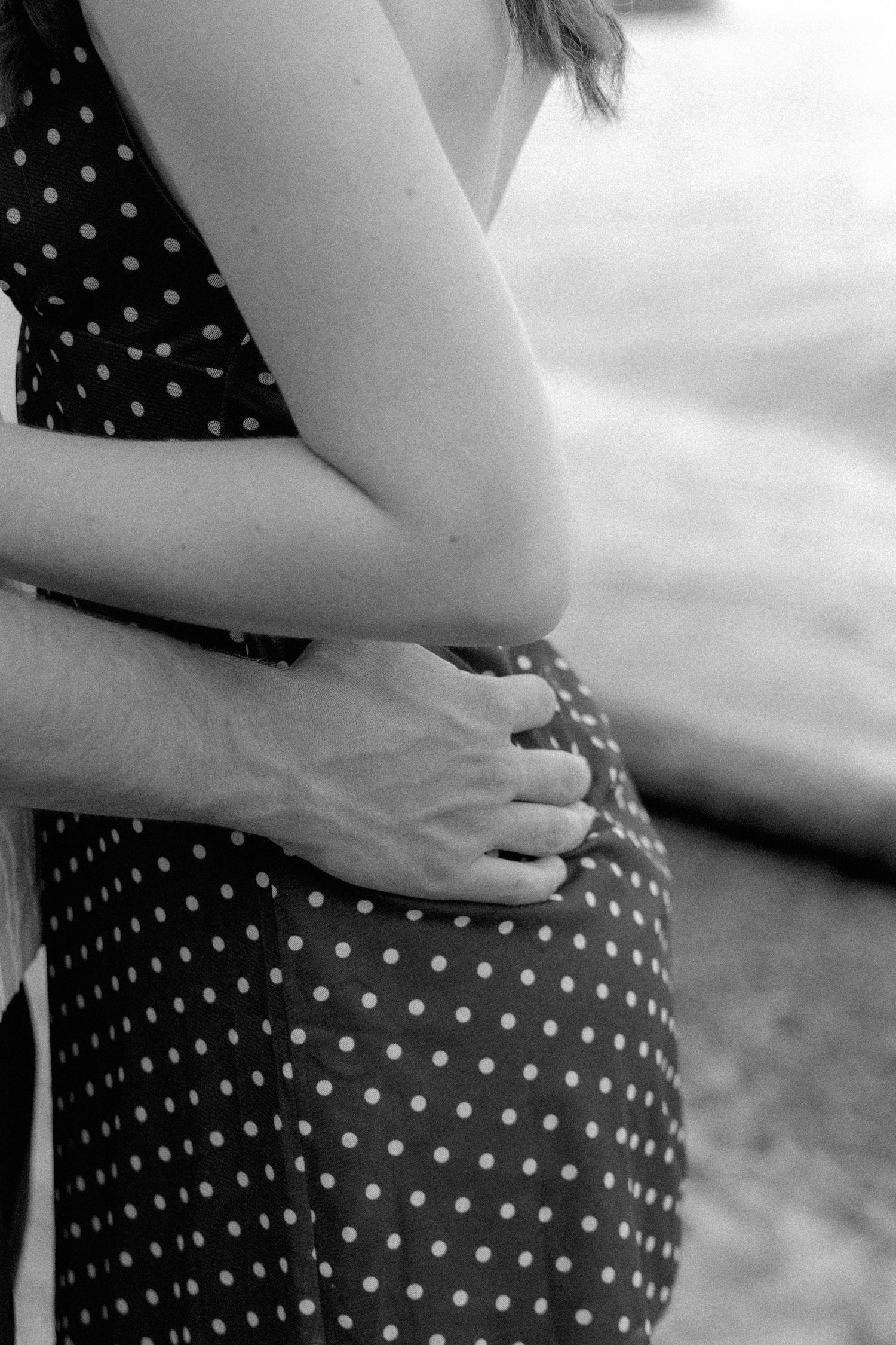 Black and white close-up of a man's hands wrapped around a woman in a polka dot dress at the beach.